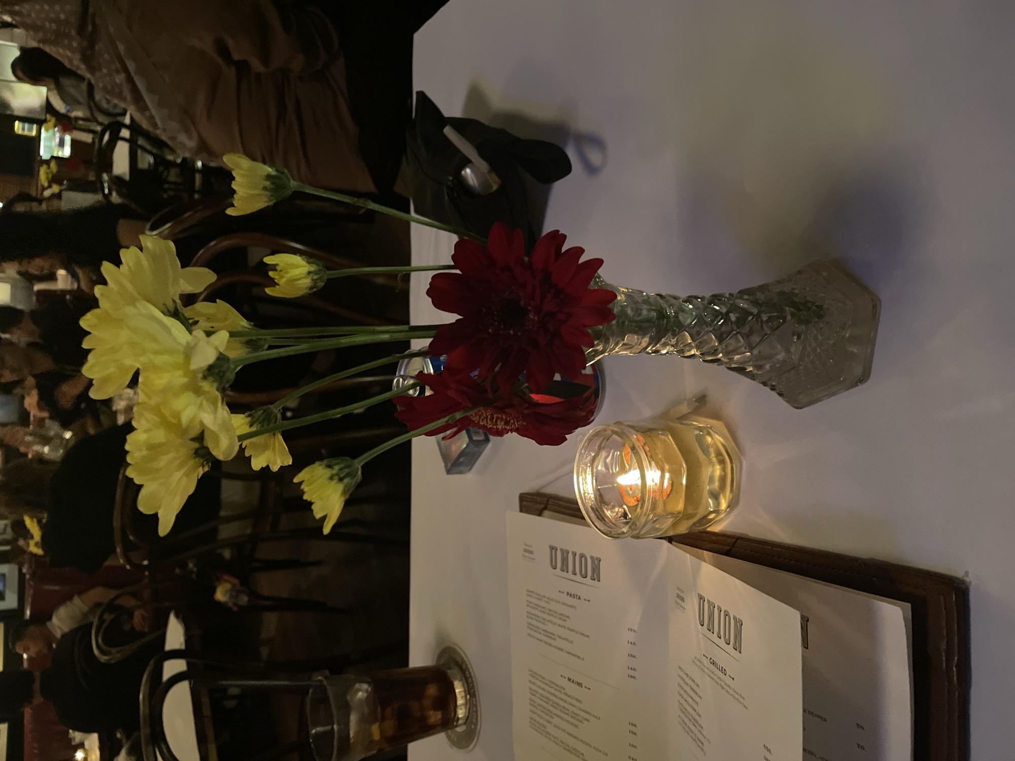 A still life on a white tablecloth features flowers, a candle, and a menu. Yellow and red flowers are arranged in a glass vase. A lit candle sits nearby. The menu, bearing the name "UNION," lists "PASTA," "MAINS," and "GRILLED" options. The scene is in Central Jakarta, Indonesia. The environment is a restaurant setting with tables and chairs visible in the background. There are people in the background.