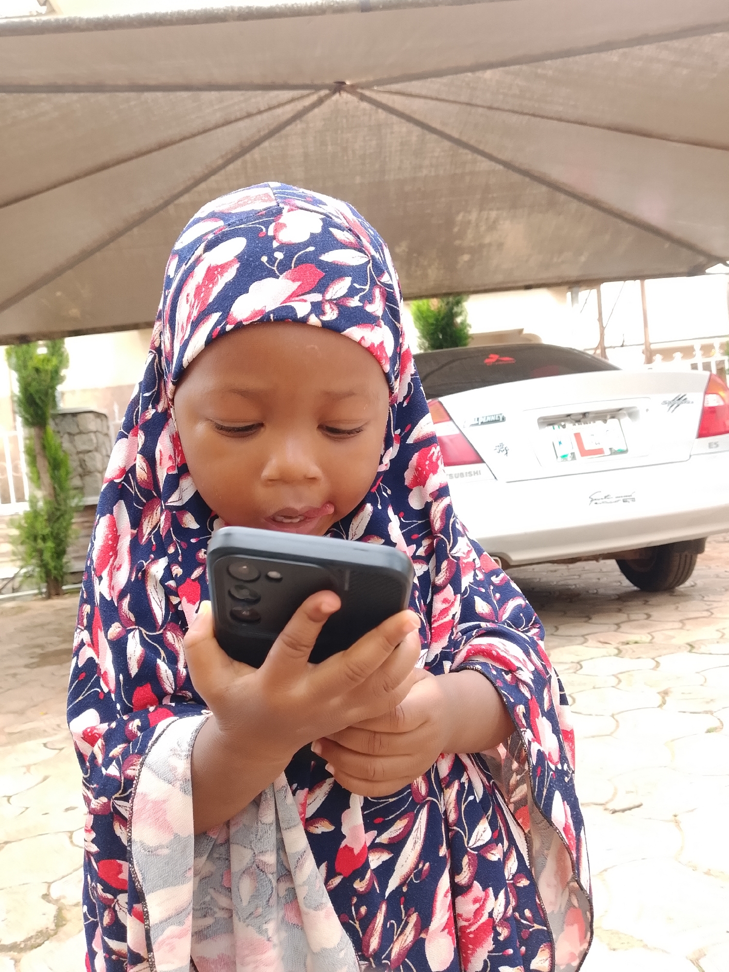In Jos, Nigeria, a young child, likely a girl given the floral headscarf and dress, is engrossed in looking at a smartphone. The child is positioned outdoors under a covered structure, possibly a carport or awning, indicated by the large fabric canopy overhead. The background features a silver Mitsubishi sedan. The ground is paved with interlocking bricks. The child is holding the phone with both hands, and their gaze is directed downwards at the screen. The overall scene suggests a casual, everyday moment captured outdoors.