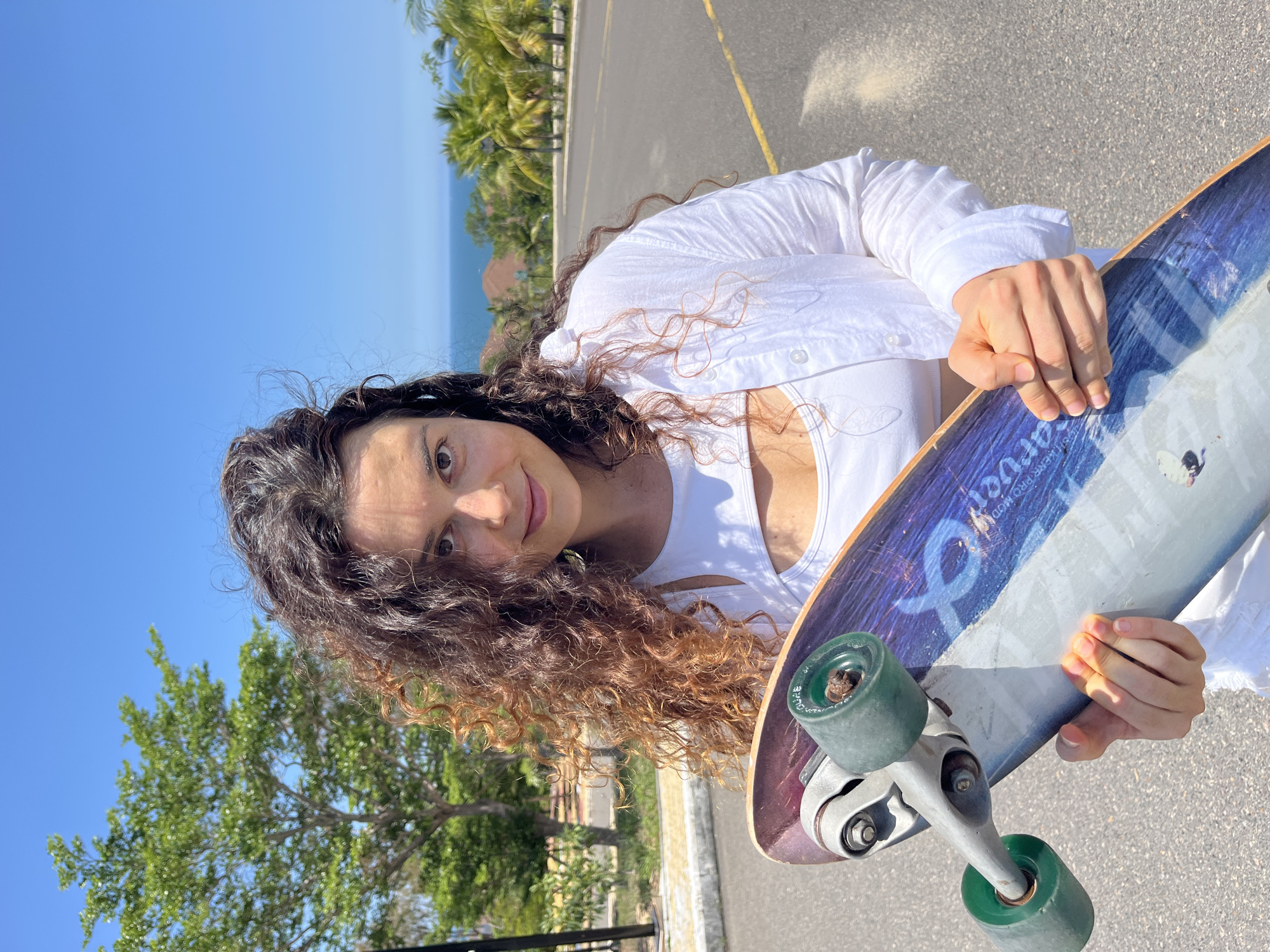 A medium shot captures a young woman with curly brown hair standing on an asphalt road in Phan Thiet, Vietnam. She is holding a skateboard with a blue and white design. She wears a white crop top and an open white button-up shirt. The background shows a tree, palm trees, and buildings, with a clear blue sky overhead and the ocean in the distance.