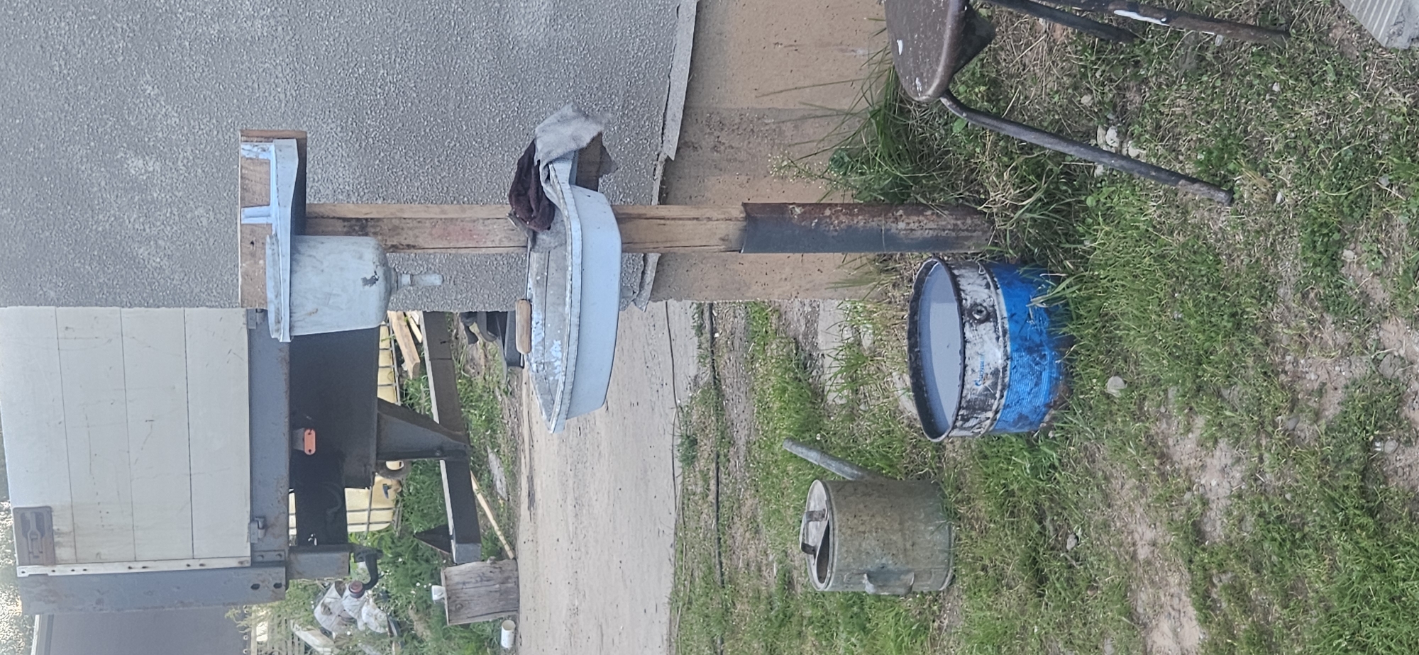 The image depicts an outdoor scene with a makeshift handwashing station. A light blue plastic basin is mounted on wooden posts, with a smaller basin above it connected to a faucet. Beside the washing station, a rusty metal watering can and a blue and white metal drum filled with water are placed on the grass. In the background, a portion of a truck or trailer with white wooden planks is visible, along with a concrete wall. The ground is a mix of dirt and grass, suggesting a rural or industrial setting. The overall impression is one of a functional, albeit rudimentary, setup for hygiene in an outdoor environment. No people are visible in the image.
