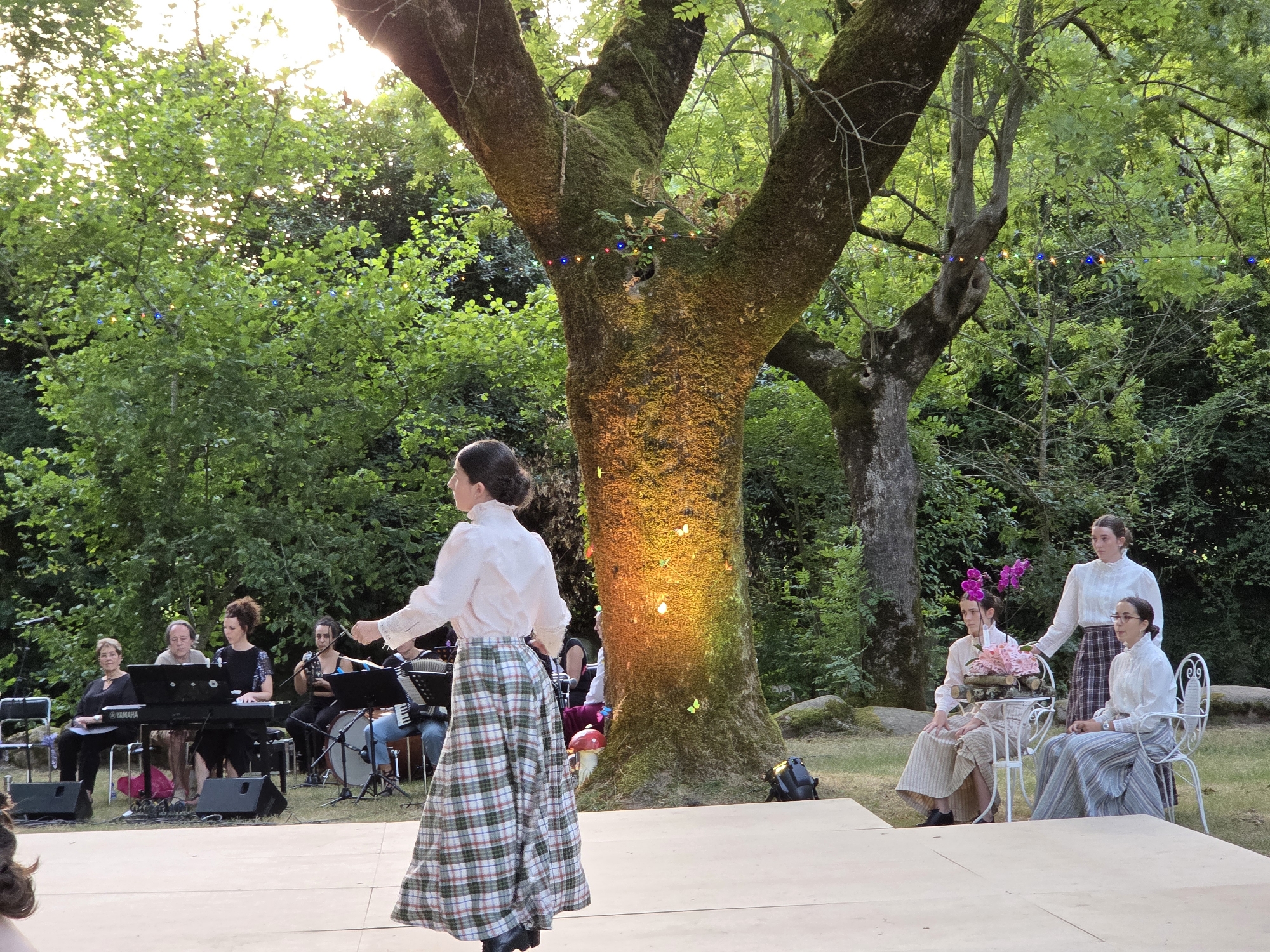 The image depicts an outdoor performance taking place in the city of Baztan, Spain. A woman in a white blouse and a long, plaid skirt stands center stage, her back to the camera, facing a group of musicians. The musicians, dressed in period attire, are seated behind keyboards, a drum, and an accordion. They appear to be playing a musical piece.
In the background, a large, moss-covered tree is illuminated by warm lights, with colorful string lights draped across its branches. The scene is surrounded by lush green foliage, suggesting a garden or park setting. To the right, three women in white blouses and long skirts are seated on chairs around a small table, adorned with a floral arrangement. The lighting suggests it is late afternoon or early evening.