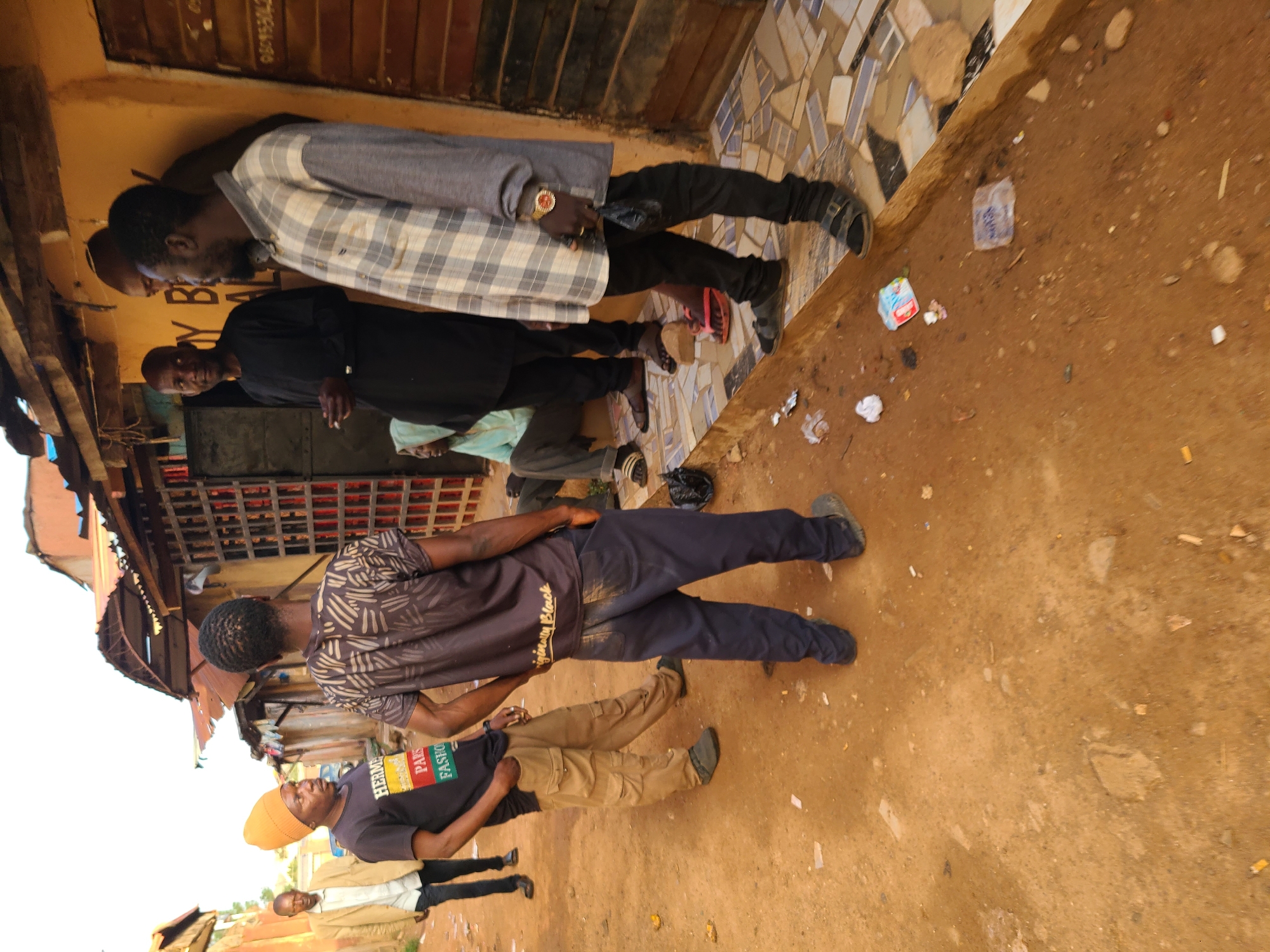 The image depicts a group of men standing outdoors in Jos, Nigeria. The ground is dirt and there is debris scattered around. The men are dressed in various casual clothing. Buildings are visible in the background. The men appear to be interacting with each other.