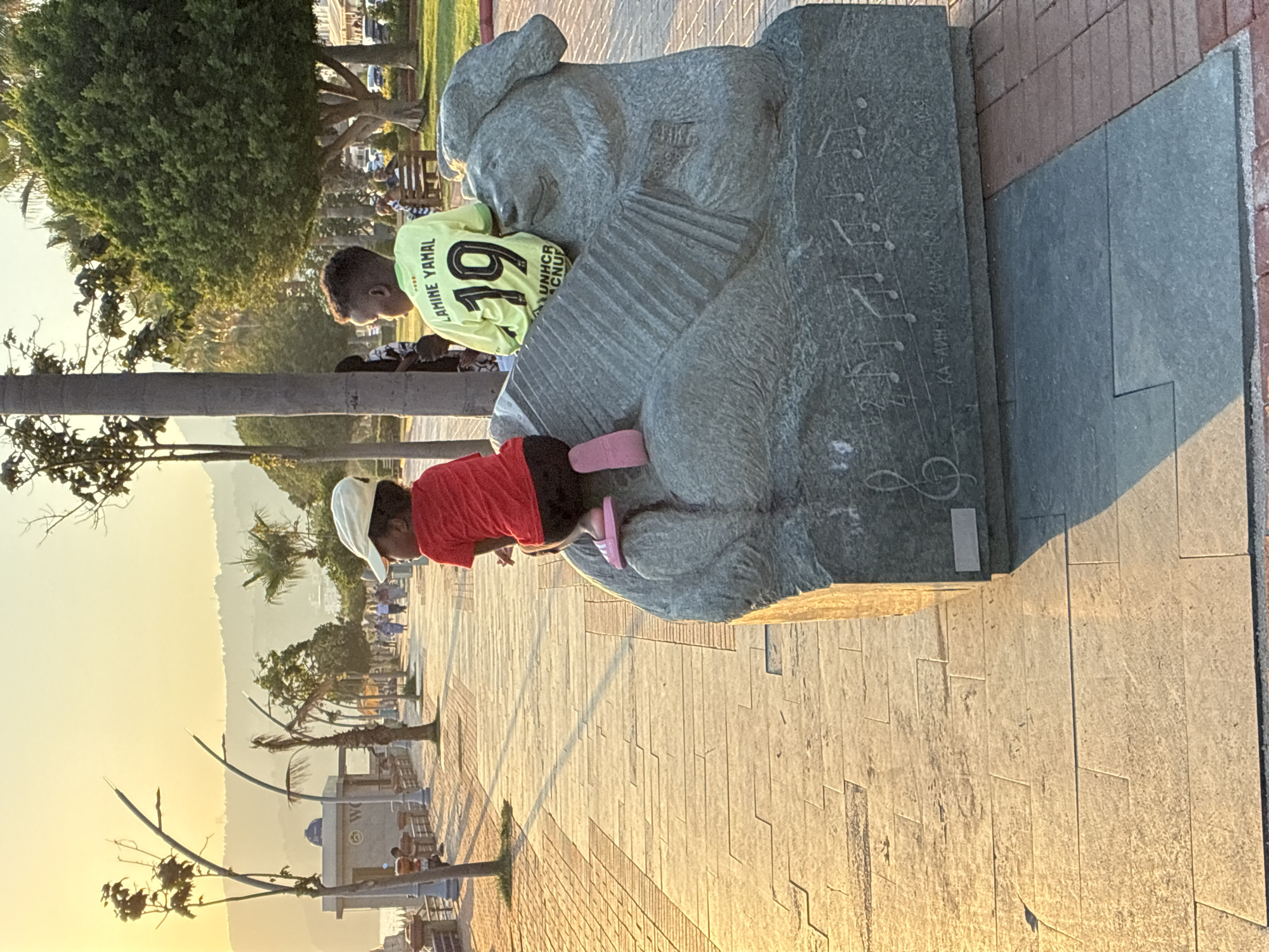 Two young boys are sitting on a stone sculpture resembling a Sphinx. The boy on the left wears a red t-shirt and black shorts, and a white cap. The boy on the right wears a lime green t-shirt with the number 19 and the name "LAMINE YAMAL" printed on the back, along with some text below. They are on a paved promenade next to a body of water, likely the Mediterranean Sea. In the background, palm trees, other trees, and buildings can be seen. The sun is setting, casting a warm glow over the scene, suggesting it is late afternoon or early evening. The sculpture is decorated with musical notes and writing. This scene is likely located in Alanya, Turkey, a popular tourist destination known for its coastal promenades and historical sites.