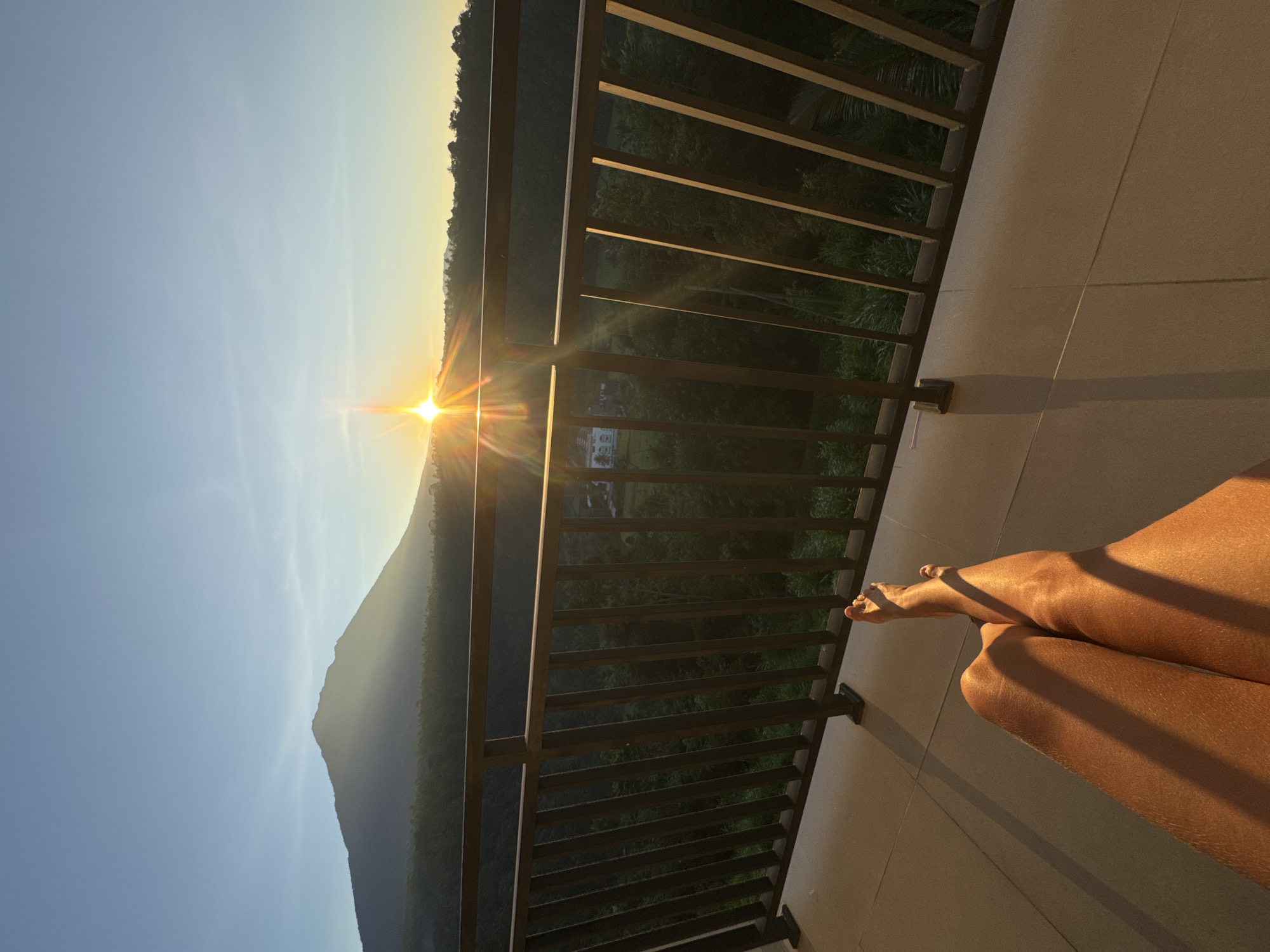 A photograph captures a moment at an elevated location, likely a balcony, in Rendang, Indonesia. The scene is bathed in the golden light of either sunrise or sunset, evidenced by the sun's placement on the horizon. A person's crossed legs extend into the frame from the right side, bearing the same warm, sunlit glow. A dark metal railing, constructed of vertical bars, separates the balcony from the view beyond. Behind the railing, a lush green landscape stretches out, possibly a hillside or forest. In the distance, a mountain silhouette is visible against the bright sky, while glimpses of buildings can be seen nestled in the greenery. The overall impression is one of relaxation and serenity, blending natural beauty with human presence.