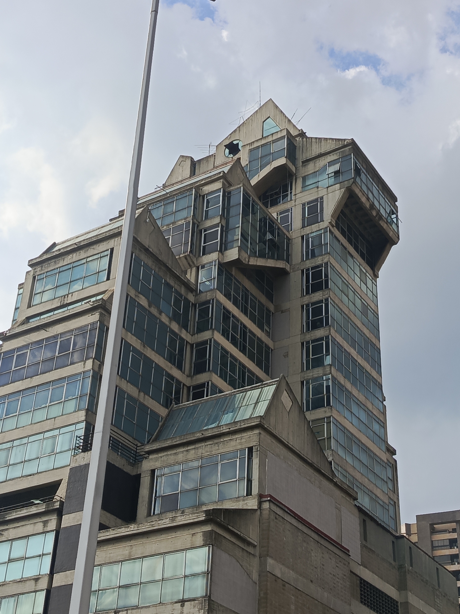 This image presents a tall, multi-story building in Caracas, Venezuela, captured on a partly cloudy day. The building's architecture is notable for its angular, brutalist-inspired concrete facade and numerous windows, many of which have a blue or green reflective tint. Some windows, particularly on the upper floors, appear open or broken, suggesting weathering or disuse.
A prominent white light pole dominates the foreground, extending vertically and partially obscuring the left side of the building. The structure itself is complex, featuring a stepped profile with various setbacks and protrusions. The uppermost section includes sharp, triangular elements and visible antennas or wires on the rooftop. Signs of aging, such as dark streaks on the concrete and wear on the materials, are evident. Lower sections incorporate different materials, possibly brick or paneling, with a visible red trim on one level. Another building is partially visible in the distant background on the right. There is no visible text or direct human activity captured in the scene. The weather is bright but with significant cloud cover.