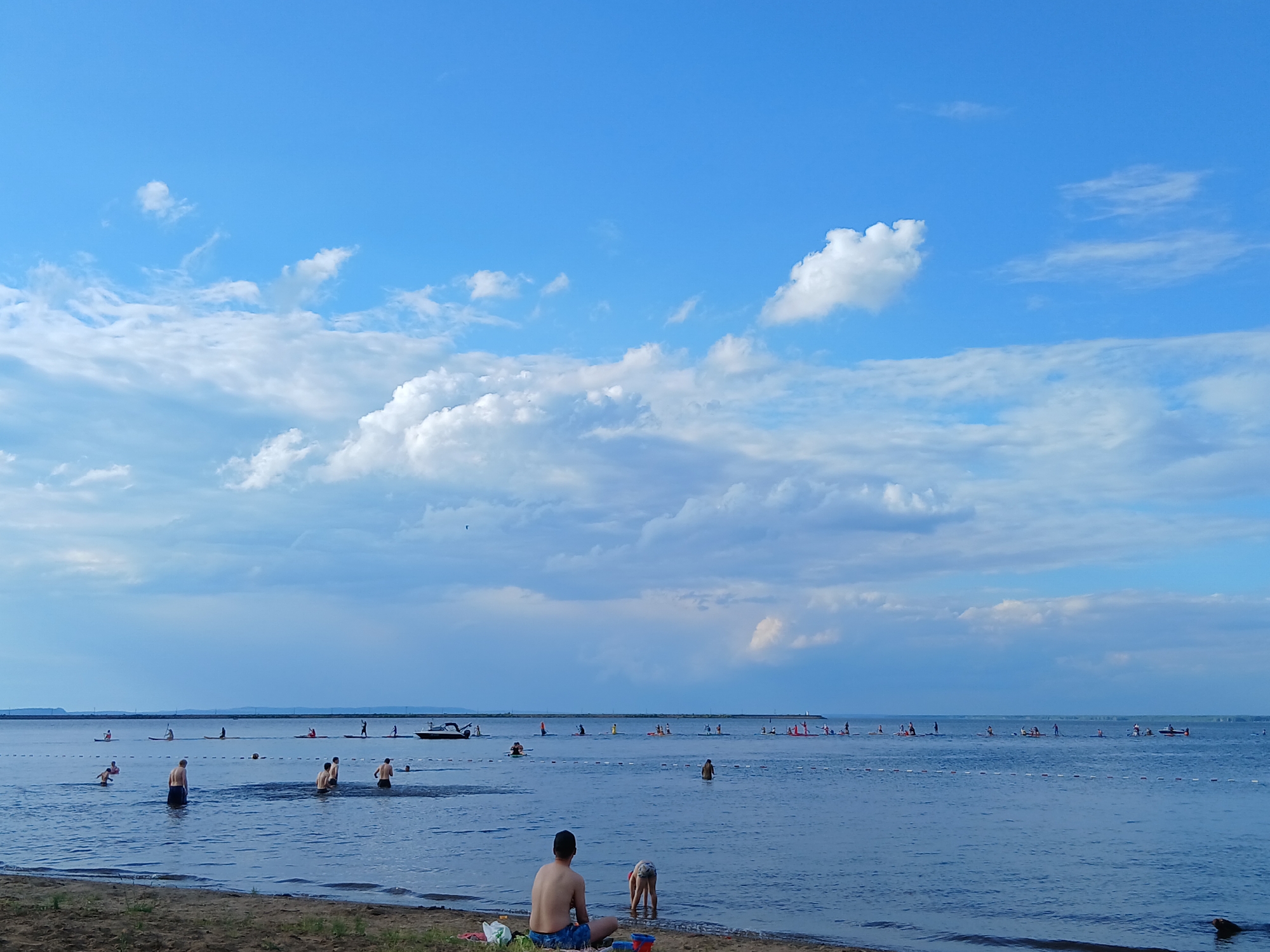 This media file captures a serene day at a beach in Chaikovsky, Russia. The scene is set against a bright blue sky dotted with fluffy white clouds, suggesting a pleasant, possibly late afternoon, time of day. The water is calm and vast, reflecting the sky.
The primary subjects are people enjoying the water and the beach. In the foreground, a shirtless man with dark hair is sitting on the sand, facing away from the camera, while a child in a swimsuit plays near the water's edge. Further out in the water, several individuals are visible. Some are swimming or wading, while a larger group is engaged in paddleboarding, spread across the expanse of the lake. A small motorboat is also present in the distance, adding to the leisurely atmosphere. The presence of a roped-off swimming area further out in the water indicates a designated recreational spot.
The overall mood conveyed is one of relaxation and summer enjoyment. The weather appears to be warm and inviting, with clear visibility and a gentle breeze suggested by the slight ripples on the water. The distant shoreline is visible as a faint line against the horizon, giving a sense of the location's geography as a large body of water. No visible text is present in the image.