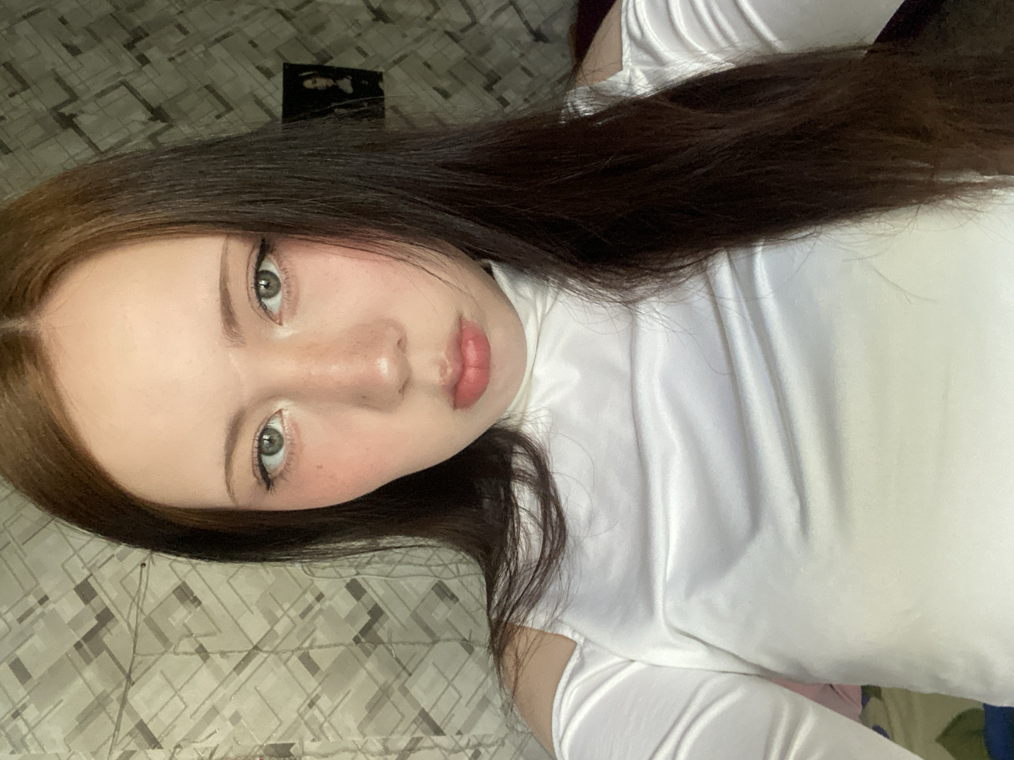A fair-skinned person with long, dark brown hair and light-colored eyes is centered in a close-up, slightly tilted portrait. The person is wearing a white, high-necked top. The background is a patterned surface, possibly a bedspread or floor covering. A small, dark rectangular object is visible in the background above the person's head. The location in the city of Unknown, Unknown is not discernible from the image.