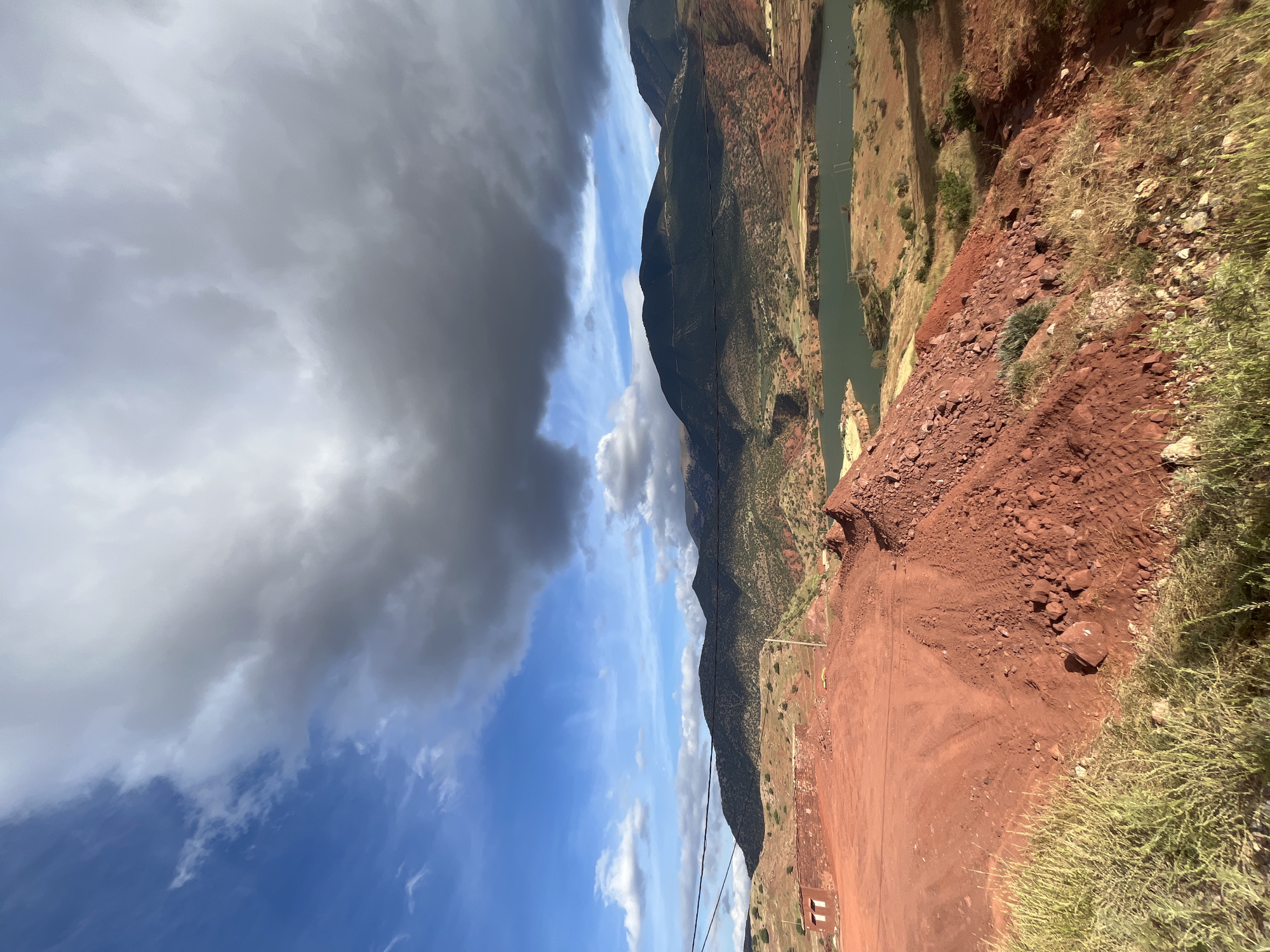 The image depicts a landscape in Ouaouizert, Morocco. A reddish-brown dirt road or path occupies the foreground, bordered by sparse vegetation. The road appears to be cut into a hillside of similar reddish-brown soil. In the background, a river flows between hills or mountains. The slopes are covered with sparse greenery and some reddish-brown areas. Overhead, the sky is partially covered by dark clouds on the left and patches of blue sky with white clouds on the right. Overhead powerlines are also visible.