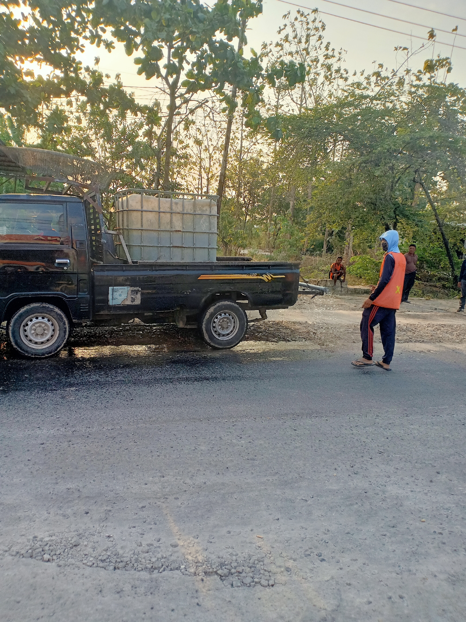 A black pickup truck is parked on a wet road, likely in Dander, Indonesia, as suggested by the presence of a water tank and construction workers. The truck bed carries a large, rectangular, translucent water tank. A man wearing an orange safety vest and a blue head covering is standing near the truck, facing away from the camera. Water appears to be spraying from the back of the truck. In the background, two other men are visible, one sitting and one standing. The scene is framed by trees and foliage under a bright, possibly early morning or late afternoon, sky. The road surface is asphalt and appears to be under repair or maintenance, with some debris present.