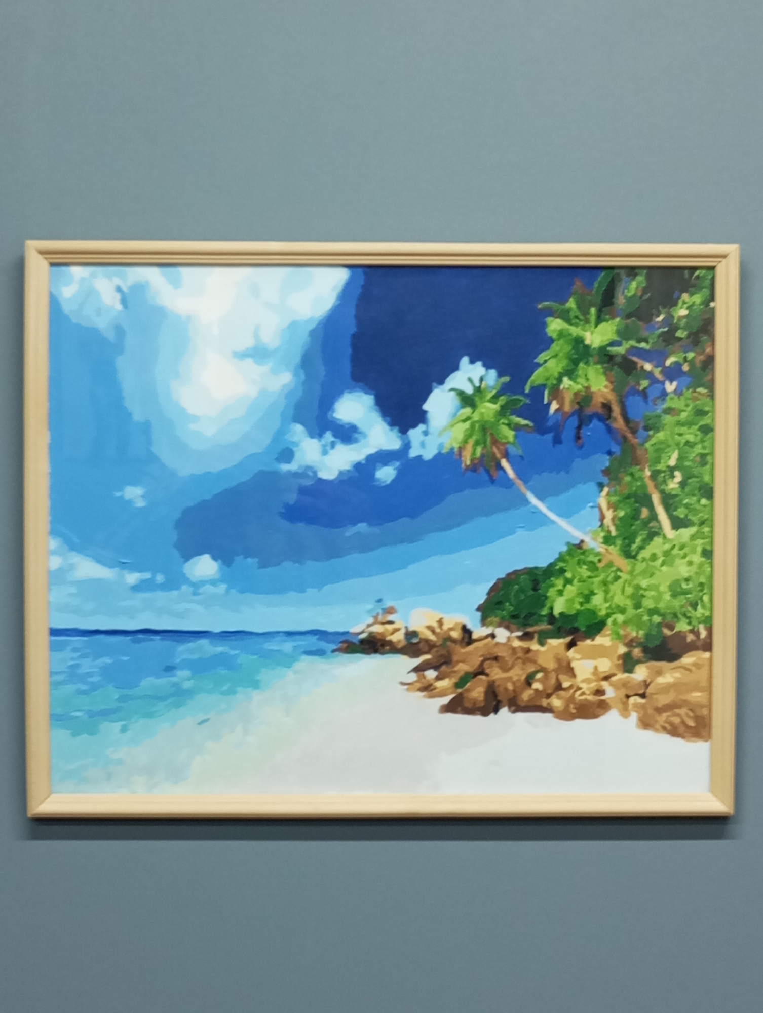 A framed painting of a tropical beach scene hangs on a light blue-gray wall. The painting depicts a white sand beach with turquoise water on the left and rocky outcroppings with green vegetation and palm trees on the right. The sky is a mix of bright blue with white clouds and dark blue patches. The scene is vibrant and colorful, evoking a sense of tropical paradise.
There is no information to verify that the location is in Pervomaysky, Russia.