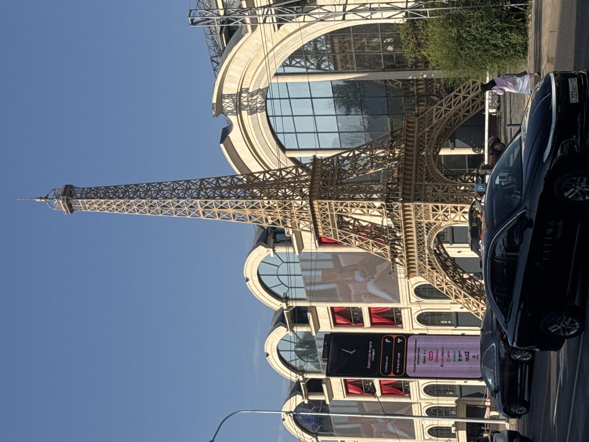 In Almaty, Kazakhstan, a prominent replica of the Eiffel Tower stands tall against a clear blue sky, casting shadows on a modern building with large arched windows and a beige facade. The tower, constructed of a lattice of golden-brown material, dominates the central part of the image. In front of the building, a tall, illuminated digital billboard displays text and logos in various colors, including "TELE2". Red curtains are visible behind the windows of the building.
The street in the foreground is paved, with several black cars driving or parked. One car in the foreground has a license plate that reads "223BD13". A woman wearing a pink top and a white skirt is walking on the sidewalk to the right of the frame, near a dense green bush. The scene appears to be a street in a commercial or entertainment district, possibly a shopping center or entertainment complex, given the presence of the large billboard and the architectural style of the building. The time of day seems to be daytime, likely late afternoon, given the angle of the sun and the shadows.