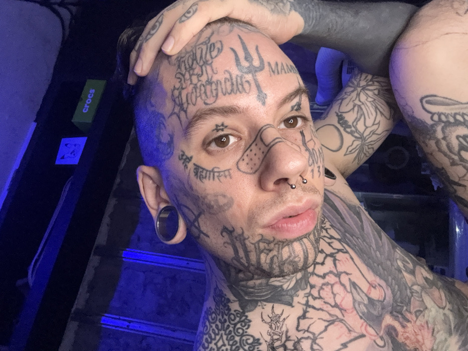 A male or masculine-presenting individual, heavily tattooed across the face, head, neck, and upper body, is depicted in a close-up, angled portrait taken indoors under blue lighting. The subject has dark, short hair, partially tinted blue, brown eyes, and full lips. Facial tattoos include text in non-Latin script (resembling Devanagari) and Latin script (e.g., 'MAMA'), a six-pointed star below the right eye, a trident-like symbol on the forehead, and an outlined cartoon band-aid on the left cheek. The individual has double nostril piercings with small silver ball studs and a large metallic ear tunnel in the left earlobe. Tattoos extending to the torso and arms include a raven/crow on the left chest/shoulder, floral patterns, abstract designs, and a blacked-out tattoo on the right arm. The subject's right hand is placed on their head, fingers resting near the right temple. The background is dark, featuring a shelf with a green 'crocs' branded object and a white object displaying a simple drawing, all illuminated by blue ambient light. The scene is set indoors in Buenos Aires, Argentina; the specific neighborhood is not discernible.