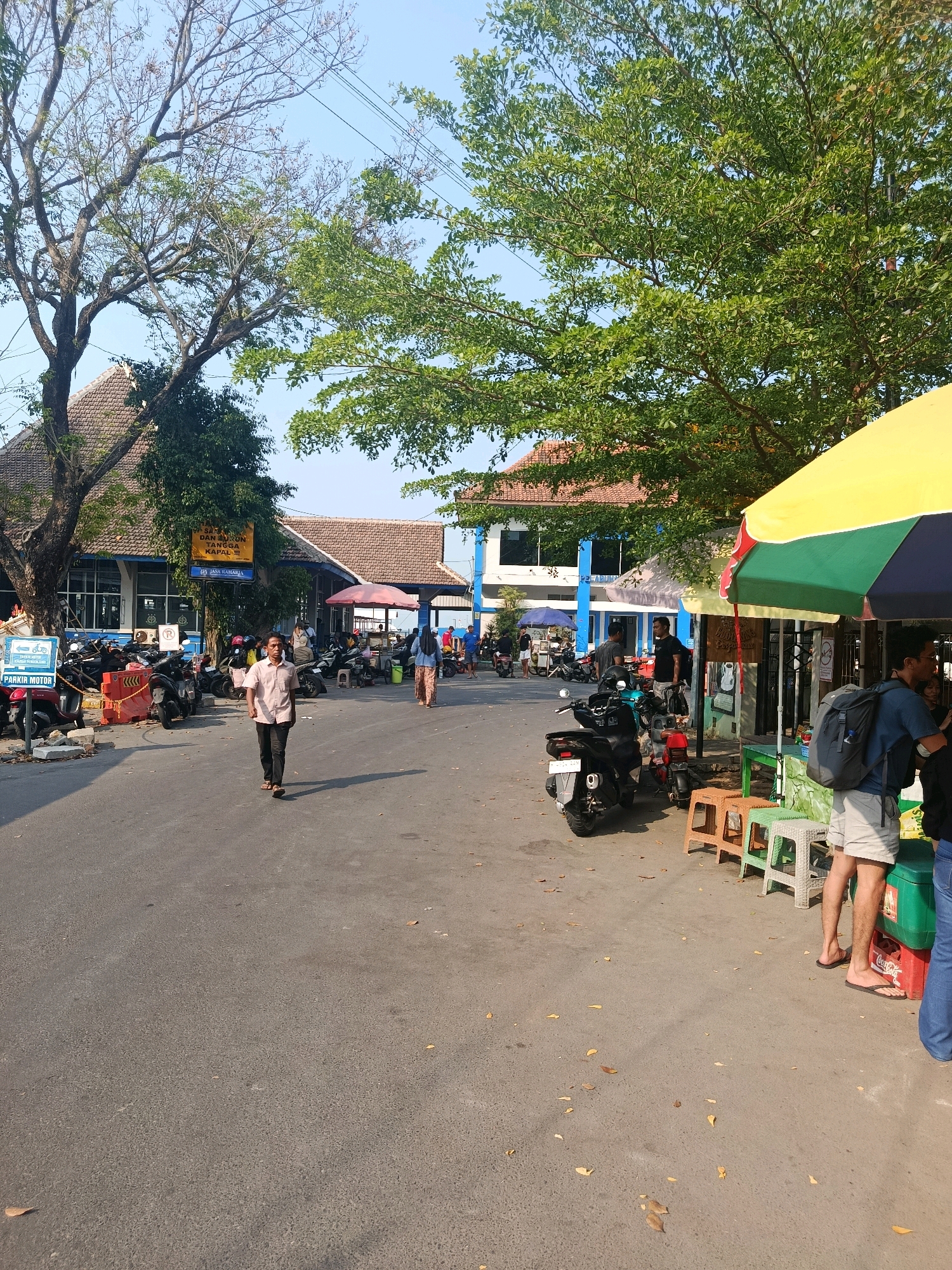 The image captures a street scene near a ferry terminal, likely in Jepara, Indonesia. In the foreground, a man in a light-colored shirt and dark pants walks away from the camera on an asphalt road. To his right, a colorful umbrella shades a small stall with several plastic stools and what appears to be a Coca-Cola cooler. Behind the stall, two men stand talking, one wearing a backpack. Further down the road, a variety of motorcycles are parked. On the left side of the road, a blue building with a tiled roof, identified by a sign as "PELABUHAN" and containing the text "JASA RAHARJA," likely indicates the ferry terminal or port authority. Another building with a similar tiled roof is visible behind it. Trees with green foliage line the street and parts of the buildings. The sky is clear and blue, suggesting a sunny day. The scene appears to be a public area with ongoing activity, typical of a transportation hub and its surrounding commercial spaces.