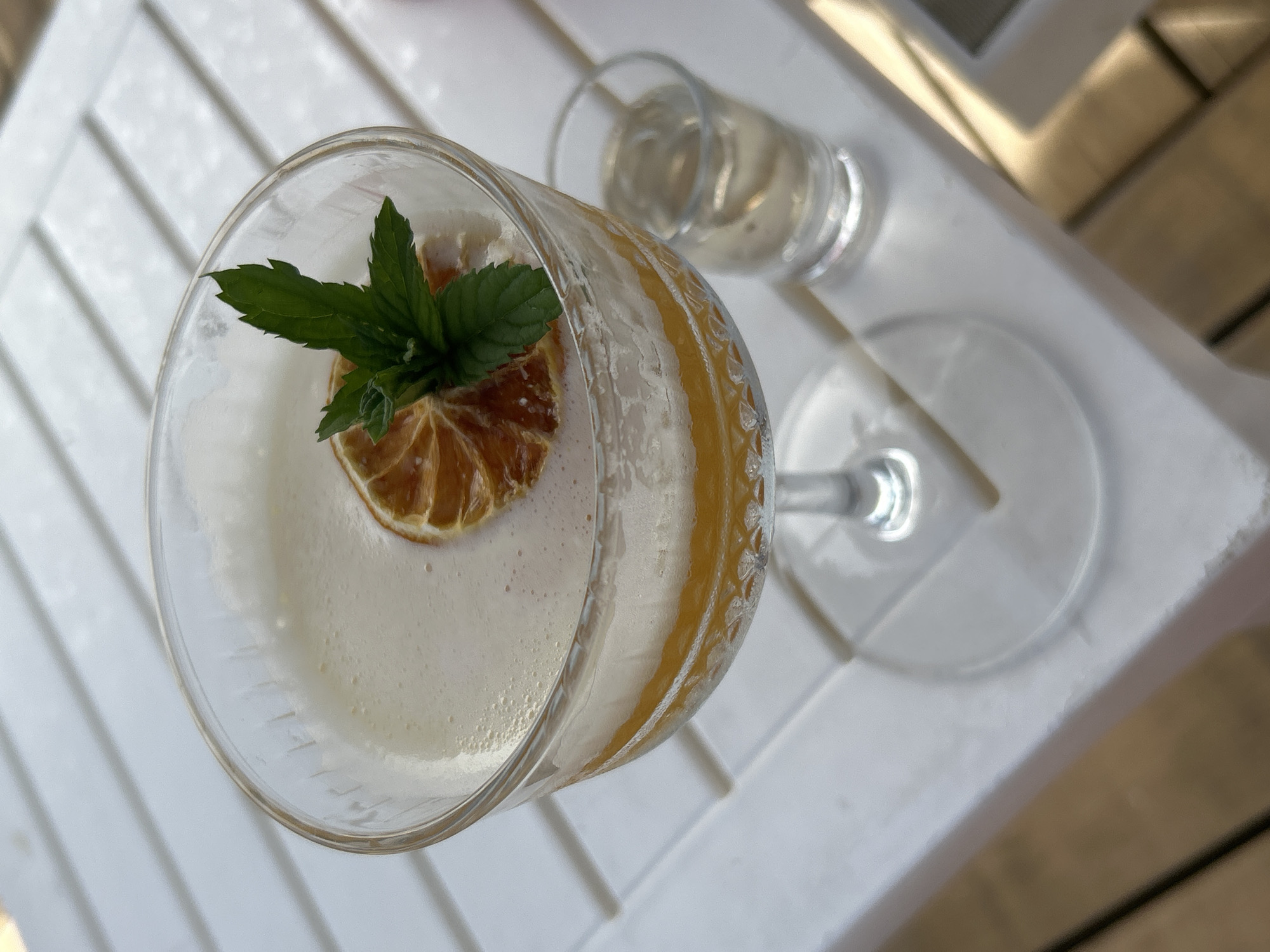 A high-angle shot shows a cocktail in a stemmed glass, garnished with a dried orange slice and fresh mint, and a small glass of liquid on a white, slatted table. The setting implies an outdoor location, possibly in Alanya, Turkey, based on prior knowledge of the image source. There are no people visible.