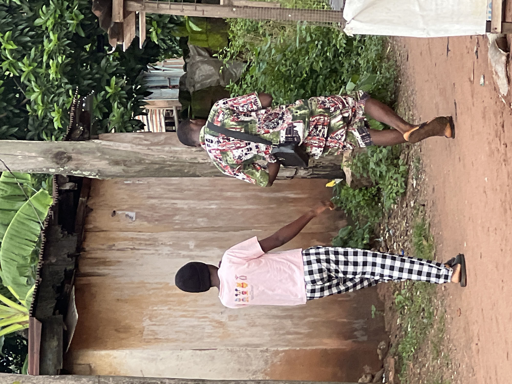 Two individuals are walking away from the camera on a dirt path in Nkpor, Nigeria. The person on the left, facing away, wears a pink t-shirt with cartoon characters and black and white checkered pants. They also wear black sandals. The person on the right, also facing away, is dressed in a patterned short-sleeved shirt and matching shorts in shades of green, red, and white. A black cross-body bag is slung over their shoulder. They wear brown sandals. The environment features a weathered, light brown building wall to the left, a wooden utility pole, dense green foliage, and the corrugated metal roof of a structure on the right. The ground is a reddish-brown dirt path with some sparse green vegetation.