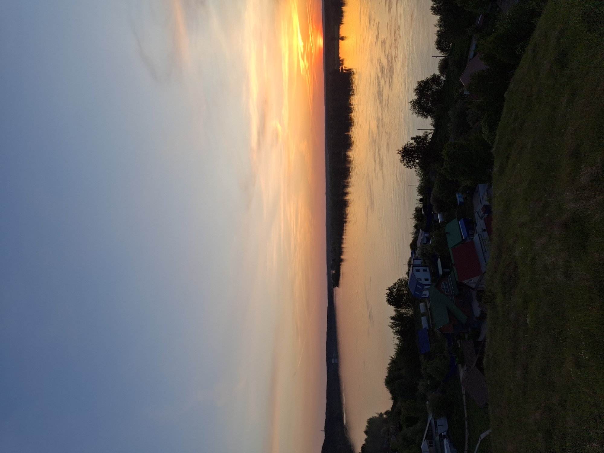 The image shows a sunset over a body of water, likely a river or large lake, located in or near the city of Ольховка, Russia. The sky transitions from blue to orange and yellow hues as the sun sets on the horizon. The setting sun reflects in the water, creating a symmetrical effect. The far side of the water body is lined with trees and a few buildings, including some residential structures with visible rooftops. A grassy hillside is present on the near side of the scene. The perspective suggests the photo was taken from an elevated position overlooking the landscape. There are no people visible in the image. The overall scene depicts a natural landscape at dusk.