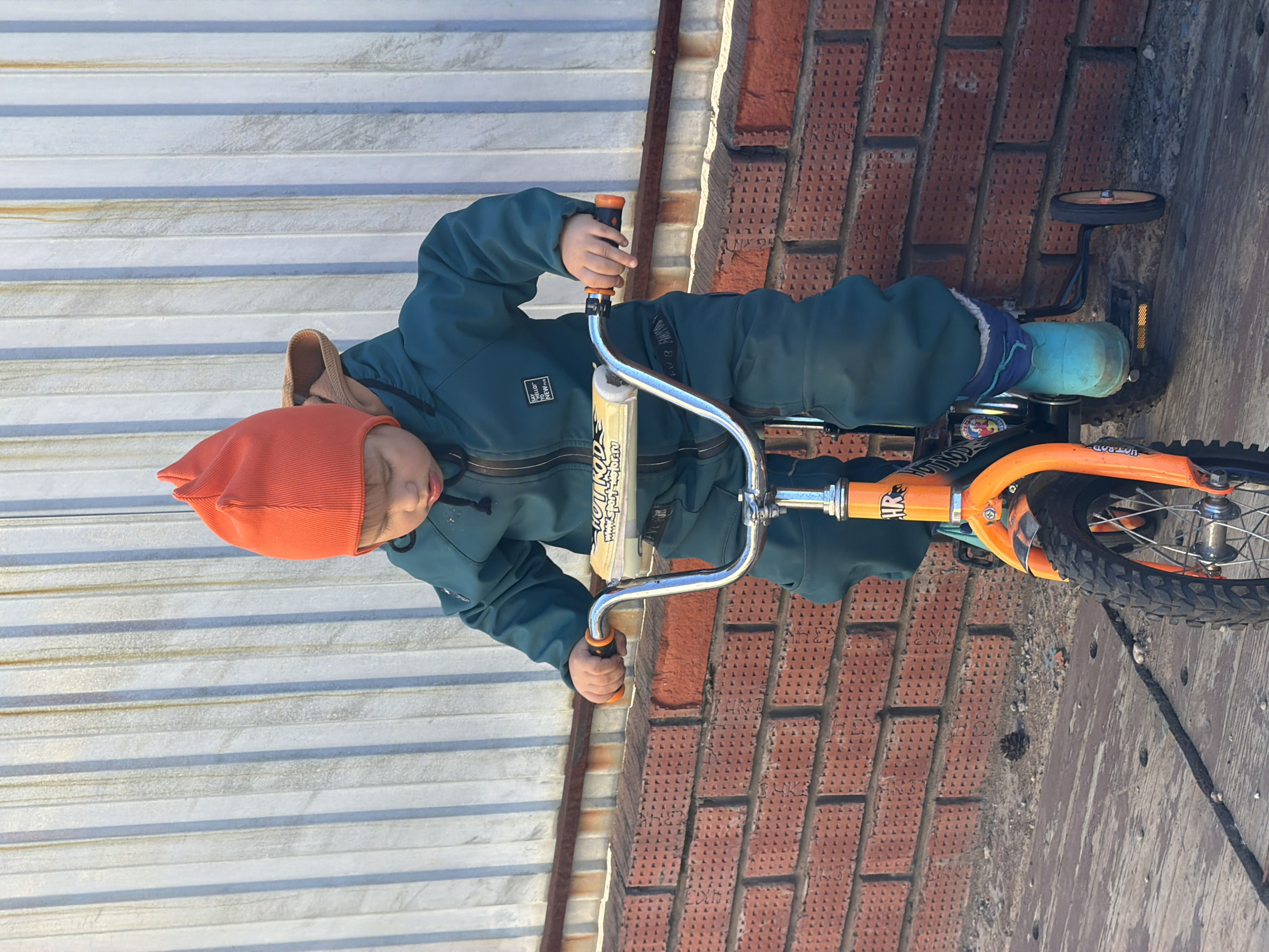 A young child wearing an orange hat and a green snowsuit is on an orange bicycle with training wheels. The child's tongue is slightly protruding. The bicycle is positioned along a brick wall and a corrugated metal wall. The ground appears to be a combination of concrete and dirt. The location is Chaikovsky, Russia.