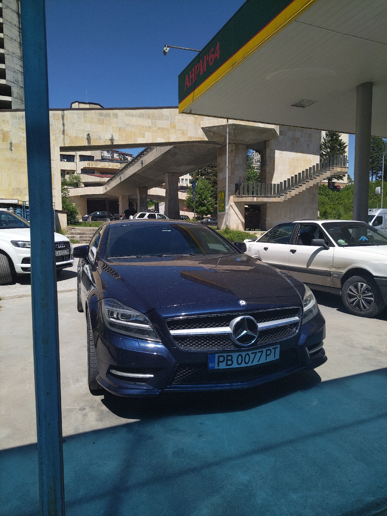The image depicts a dark blue Mercedes-Benz CLS with license plate "PB 0077 PT" parked at a petrol station in Smolyan, Bulgaria. A white Audi SUV with license plate "CA 4225 TB" and a white Audi sedan are parked nearby. The petrol station has a sign with the text "АНРИ '64". In the background, a multi-story concrete building with a pedestrian overpass and outdoor stairs are visible. The scene is set on a sunny day.