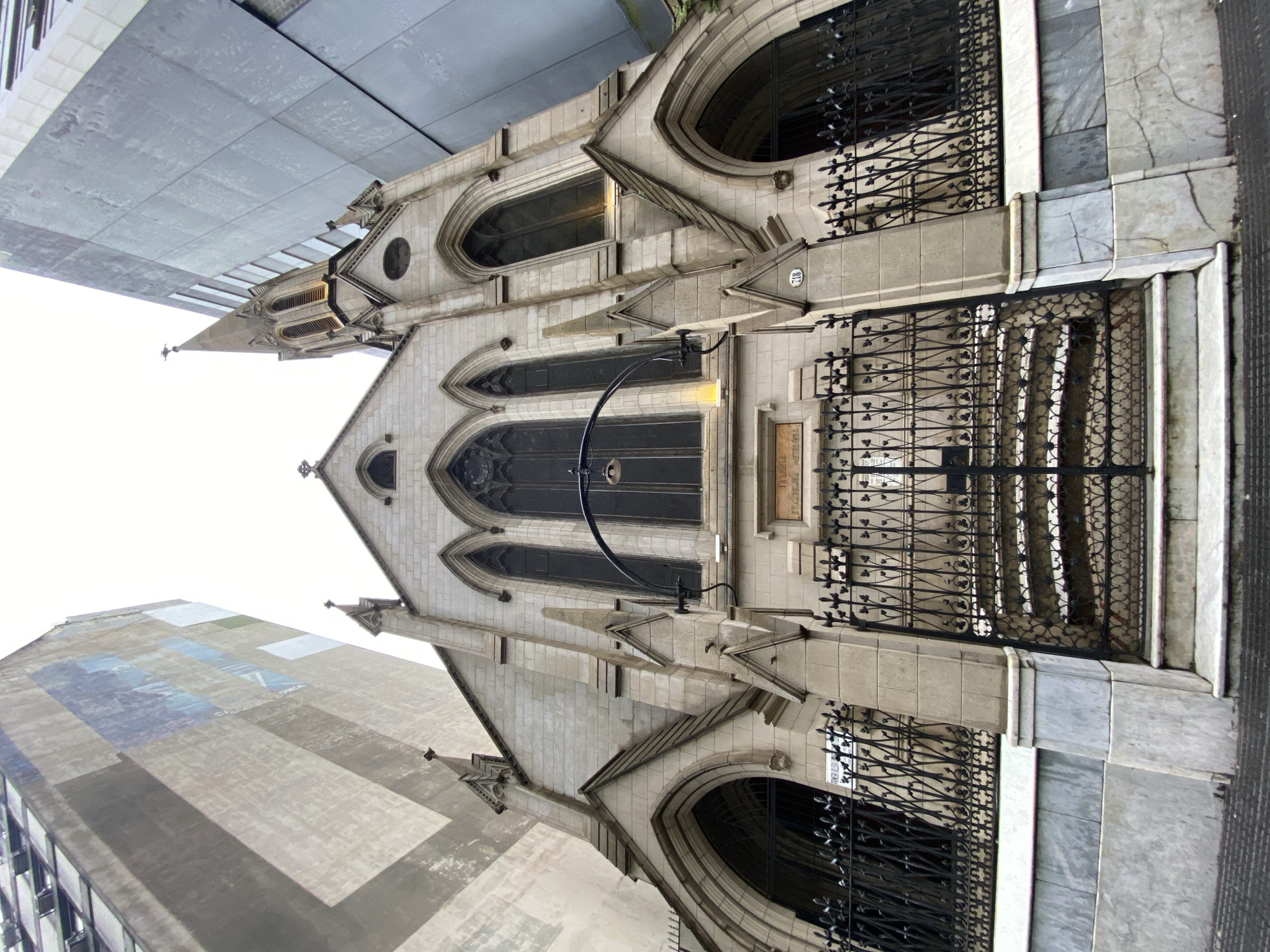 The image shows the facade of the Iglesia Dinamarquesa de Buenos Aires, a Danish church located in Buenos Aires, Argentina. The church features Gothic Revival architecture with pointed arches, ornate windows, and decorative ironwork. The building is made of light-colored stone. There is a sign visible near the entrance, with the church's name. Above and beside the church, modern high-rise buildings loom, illustrating a contrast between historical architecture and contemporary urban development. The photo is taken from a low angle, emphasizing the church's height relative to the viewer. The sky is overcast.