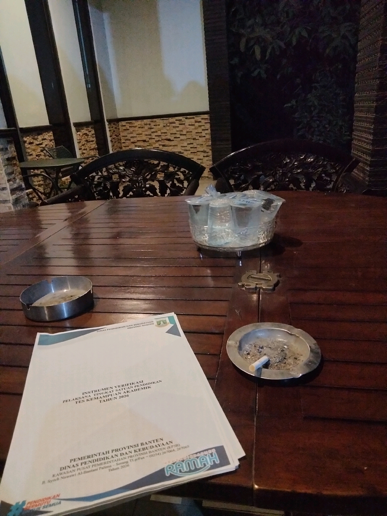 A low-angle, indoor photograph taken at night shows a wooden table with various objects on it. The table appears to be made of dark, polished wood with a visible wood grain pattern. In the foreground, a white document with text and a seal is partially visible on the left. To the right of the document, a metal ashtray contains cigarette butts and ash. Further back on the table, a clear plastic container holds several clear plastic cups filled with a clear liquid. The background shows a dimly lit area with decorative chairs and a brick-patterned wall. The scene suggests a casual or informal setting, possibly a restaurant or cafe, in Suka Sari, Indonesia.