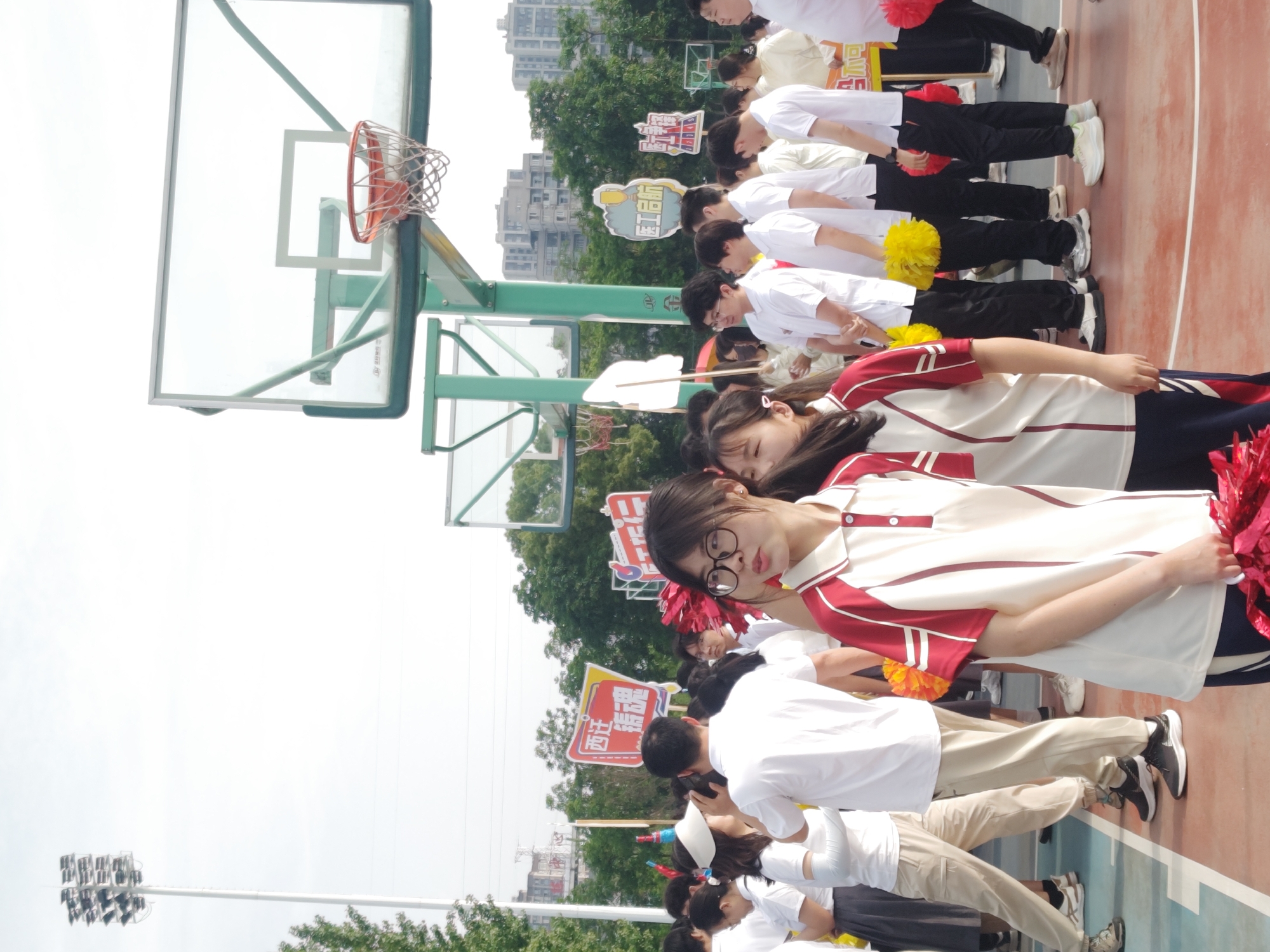 A line of students stand on an outdoor basketball court in the Chongqing High-tech Zone, China. The students are dressed in uniforms and hold pom-poms and signs. A basketball hoop is in the background, along with trees and buildings. Overhead lighting is visible in the top-left corner.