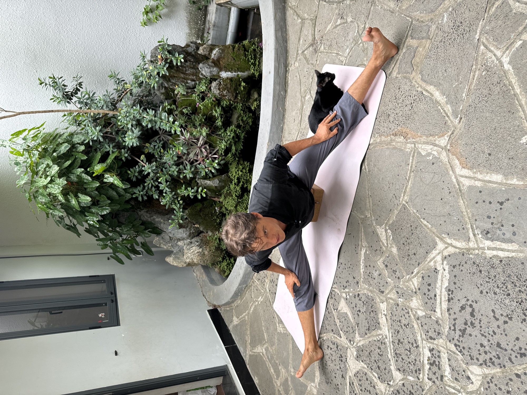 A man is performing a yoga pose outside in Mũi Né, Vietnam. He is wearing grey pants and a black shirt and is sitting on a pink yoga mat on a stone tile patio. He is using a yoga block under his hips. A black cat is sitting on his legs. There is a decorative wall and ledge with plants nearby, and a window is visible on the wall behind him.