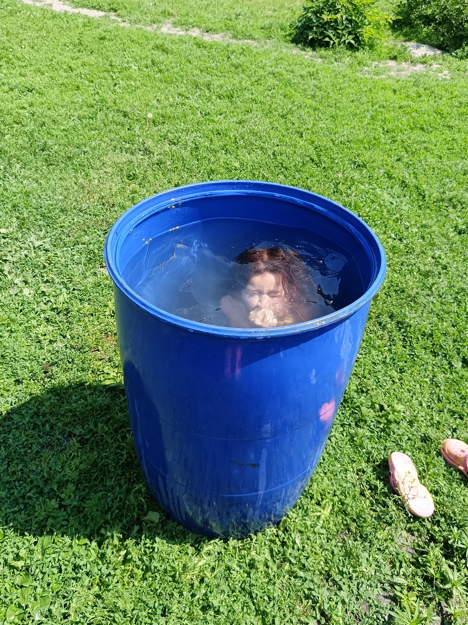 A young person is submerged in a large blue plastic barrel filled with water, with their face partially visible above the surface. They are holding something in their hands near their mouth. The barrel is situated outdoors on a grassy area, with green grass covering the ground. In the background, there are bushes and a dirt path. Two pink flip-flops are lying on the grass to the right of the barrel. The scene appears to be in a rural setting, potentially a backyard or a dacha, given the natural environment and the presence of basic amenities like the barrel and flip-flops. The lighting suggests it is daytime, with sunlight casting shadows. The location is specified as Lagovskoe, Russia.