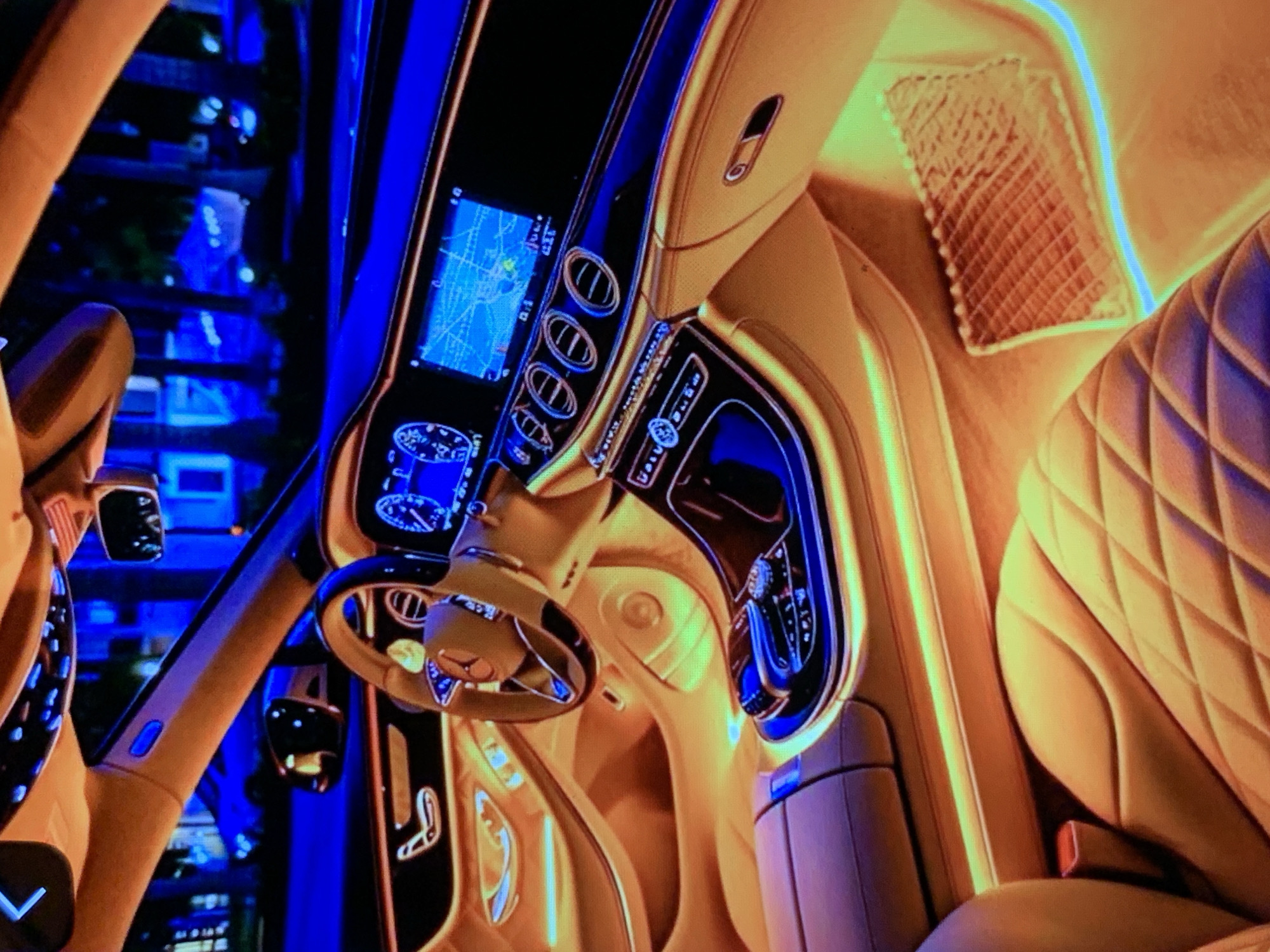 The image shows the interior of a luxury car at night. The car is a Mercedes-Benz, as indicated by the logo on the steering wheel. The interior is illuminated by ambient lighting in shades of orange, yellow, and blue, which creates a dramatic and luxurious atmosphere. The seats are made of light-colored leather with a diamond-stitched pattern. The dashboard features two large screens, one displaying navigation information and the other showing car diagnostics. The center console has several buttons and a rotary dial. The exterior of the car is visible through the windows, with streetlights and trees suggesting an urban or suburban setting. The overall impression is one of comfort, technology, and sophistication.