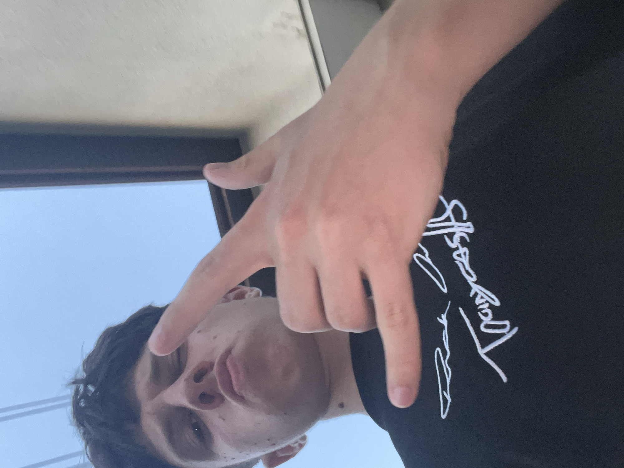 A low-angle self-portrait features a young male subject with dark hair, light skin, and visible moles on his cheeks and neck. His eyes are partially closed, and his lips are pursed as he looks directly at the camera. The subject's left hand is raised in the foreground, performing a "rock on" gesture with the index and pinky fingers extended. He is wearing a dark t-shirt, possibly black, adorned with white cursive text on the chest, which includes an inscription resembling "Trapycas" and another less discernible signature above it. The background consists of a clear light blue sky in the upper left, transitioning to a dark-colored structural element, possibly a window frame, and a light-colored, textured building wall on the right side. The image contains no discernible landmarks or contextual information to confirm a specific location within Zlatograd, Bulgaria.