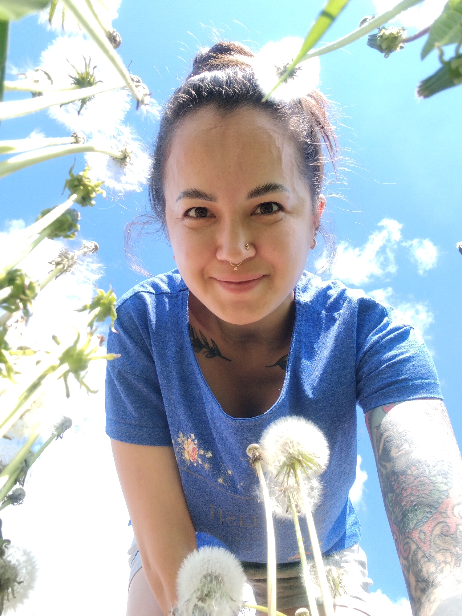 A woman with dark hair pulled into a bun, wearing a blue t-shirt, is holding dandelions and looking directly at the camera from a low angle. She has a nose ring, earrings, and tattoos on her neck and left arm. The background is a bright blue sky with scattered clouds, visible through the dandelions. The location is not specified, and there is no indication that the image was taken in Гожан, Russia.