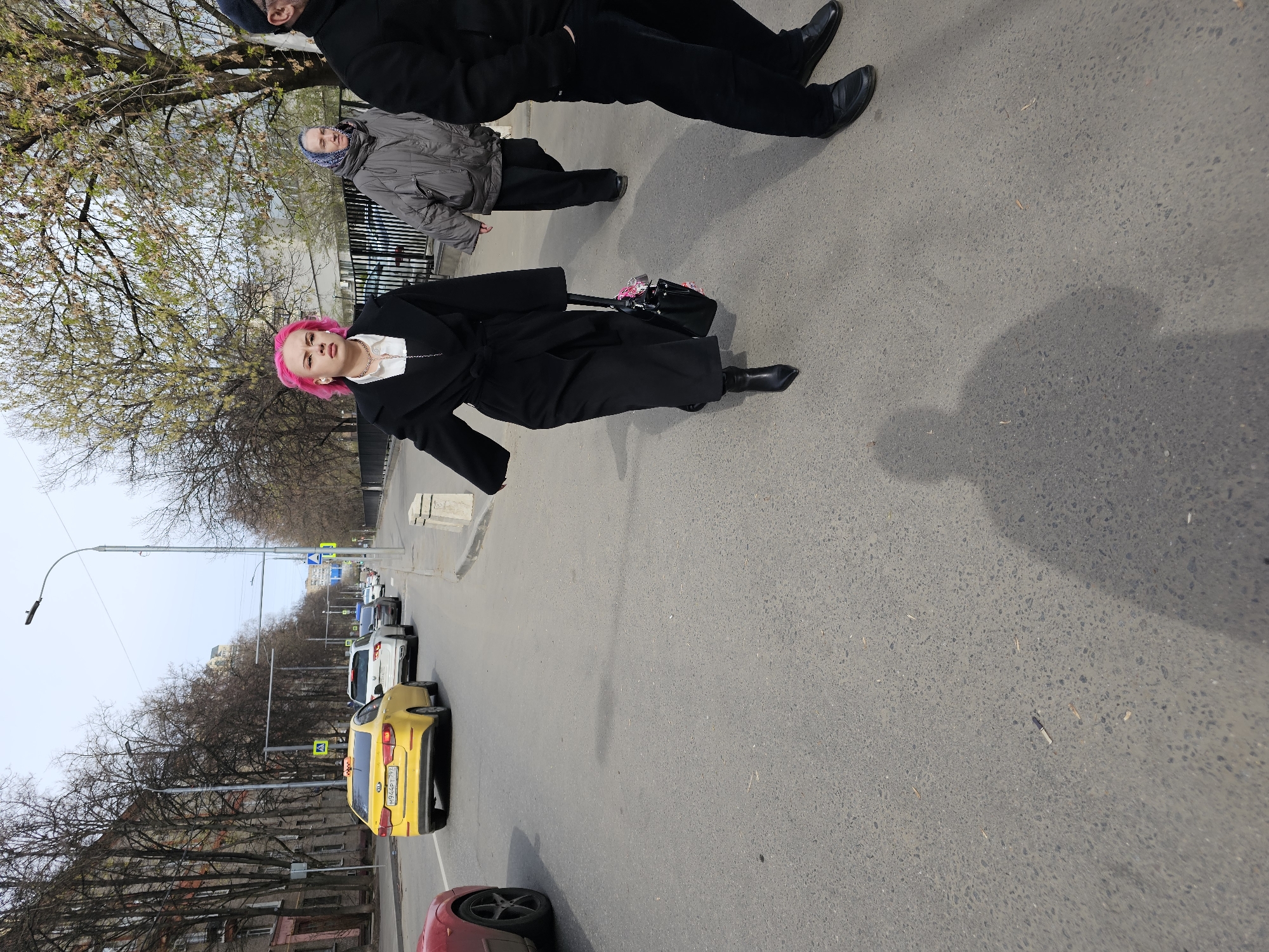 The image captures an outdoor urban scene, rotated ninety degrees clockwise, showing people on a sidewalk adjacent to a street in what appears to be Khimki, Russia. The sky is overcast, and the trees lining the street are largely bare or show early spring buds, suggesting cool weather during daytime.
The central subject is a young person with vibrant bright pink hair, wearing a long black coat over a white top with a gold-colored chain, black pointed-toe boots, and white wireless earbuds. They hold a black bag with pink accents and look directly at the camera with a neutral expression.
Behind this individual, slightly to the left, stands an older person in a gray puffer jacket and a blue patterned head covering. Further to the right and partially visible are two other individuals, mostly seen from the waist down, dressed in dark clothing and black shoes.
The asphalt road features a prominent yellow Kia Rio taxi with the license plate "M914PP 799" (Moscow region, Russia) visible on its rear. Other cars are parked or moving in the distance. The street is equipped with streetlights and concrete barriers. In the background, multi-story buildings line the street. A discarded cigarette butt is visible on the asphalt.
The scene depicts people walking casually in an urban environment, with the pink-haired individual appearing to acknowledge the camera. The striking hair color of the central subject is a notable detail.