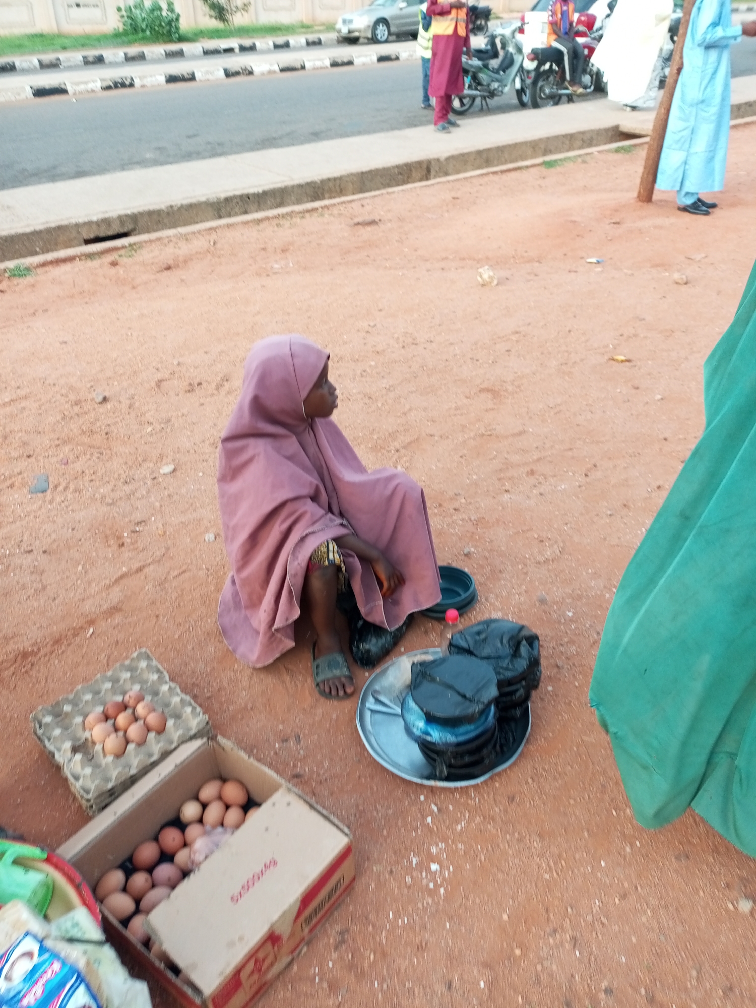 The image depicts a street scene in Gombe, Nigeria, with a young girl wearing a mauve hijab and traditional clothing sitting on the dusty ground. She is surrounded by items she appears to be selling, including several cartons of eggs. To her right, a large green cloth hangs from an unseen structure, partially obscuring the background. In the distance, several people are visible near motorcycles and a white car, suggesting a busy area. The road and sidewalk are visible in the background, indicating the setting is likely a roadside market or a place where vendors gather. The ground is composed of reddish-brown dirt and small stones.