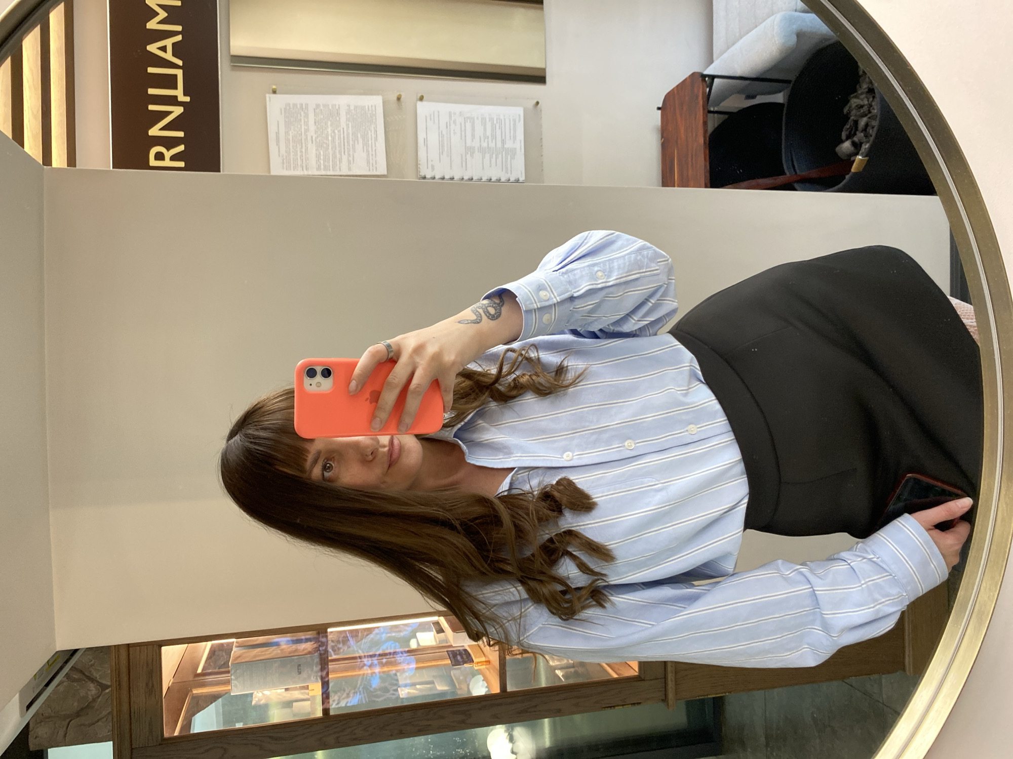 A woman with shoulder-length brown hair and bangs is taking a selfie in a mirror. She is wearing a light blue striped, long-sleeved button-down shirt and a black skirt. She holds an orange smartphone in her left hand. The reflection suggests she is inside a shop, potentially in Votkinsk, Russia, based on the Russian text "RNLIAИ" visible on a sign in the upper left corner of the frame. Behind her is a display case and a wall with documents hanging on it. In the upper right is a glimpse of a decorative bowl or container.