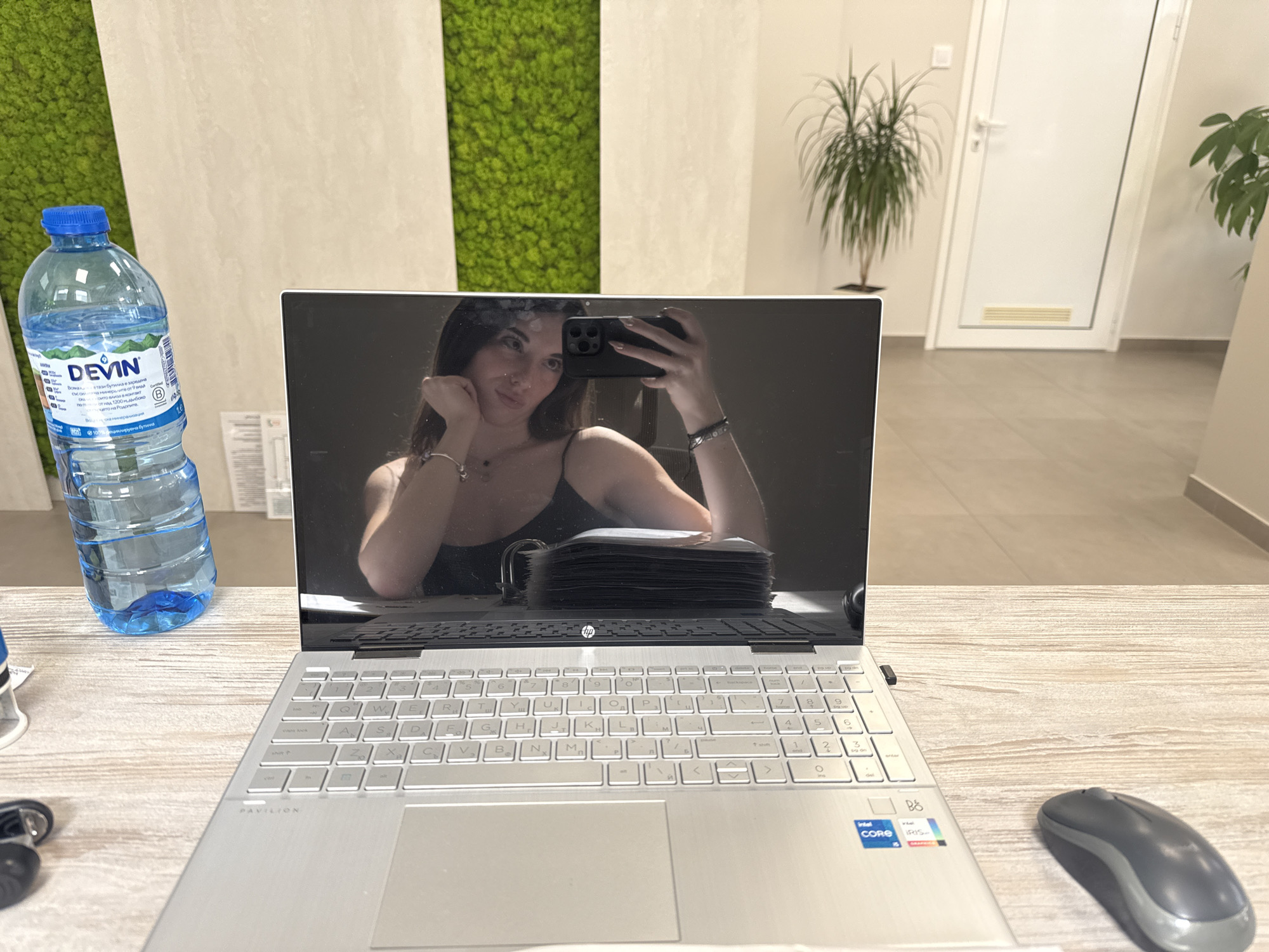 A young woman is taking a selfie in the reflection of a laptop screen. She is leaning her head on her hand, with her arm resting on a stack of papers. She is wearing a black tank top and a necklace. The laptop is open and placed on a wooden desk. Next to the laptop is a bottle of water and a computer mouse. In the background, there is a wall with green moss panels, a plant, and a white door. The scene is likely in an office or a modern workspace in Trud, Bulgaria, suggested by the presence of the DEVIN water bottle, a popular Bulgarian brand.
