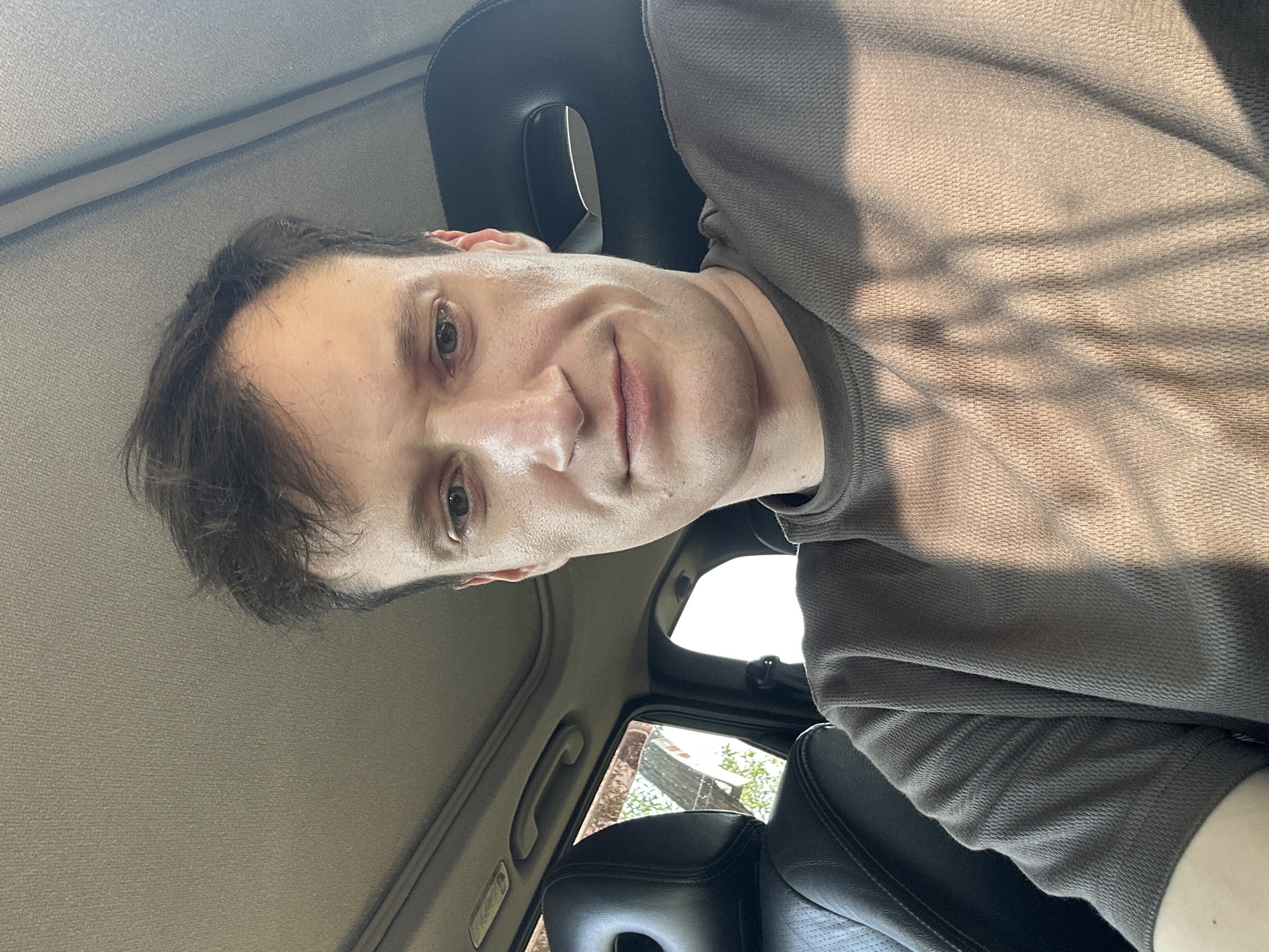 A young, fair-skinned man with short, dark brown hair is seated in the driver's seat of a car. He is looking directly at the camera with a slight smile. He is wearing a grey, short-sleeved t-shirt. The interior of the car is visible, including the dark leather seats and the sunroof. Sunlight is filtering into the car, casting shadows on the man's shirt and face. The scene appears to be set in Makiv, Ukraine, as indicated by the information provided.