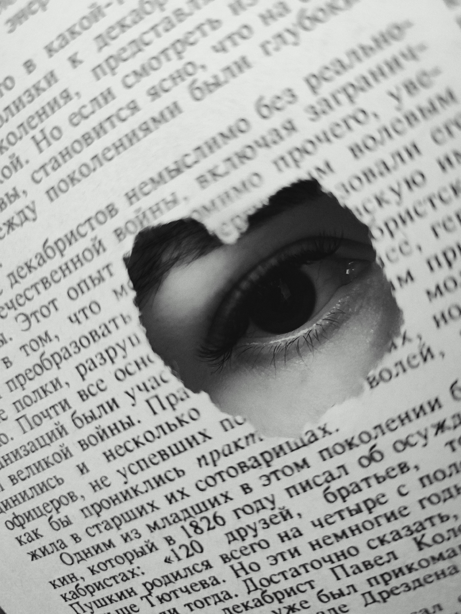 The image is a close-up shot of a book page with a hole torn through it, revealing a human eye looking through the gap. The book is open, and the text is in Russian. The text is about the Decembrists, a group of Russian revolutionaries. The eye is focused and appears to be staring directly at the viewer. The overall effect is both mysterious and unsettling. There is no information about a location in the city of Fokino, Russia, in the image.