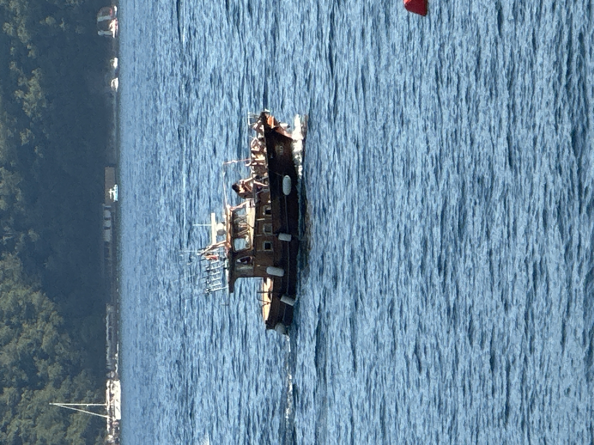 A wooden boat with several people on board is moving through blue water. The boat is brown and has a cabin, fishing rods, and several buoys attached to its sides. Some of the people on the boat appear to be sunbathing or relaxing. In the background, there is a lush green forested hillside and several other boats, including a sailboat, and some buildings along the shore. The overall scene suggests a sunny day in a coastal area, possibly Budva, Montenegro, given the presence of the characteristic blue water and greenery.