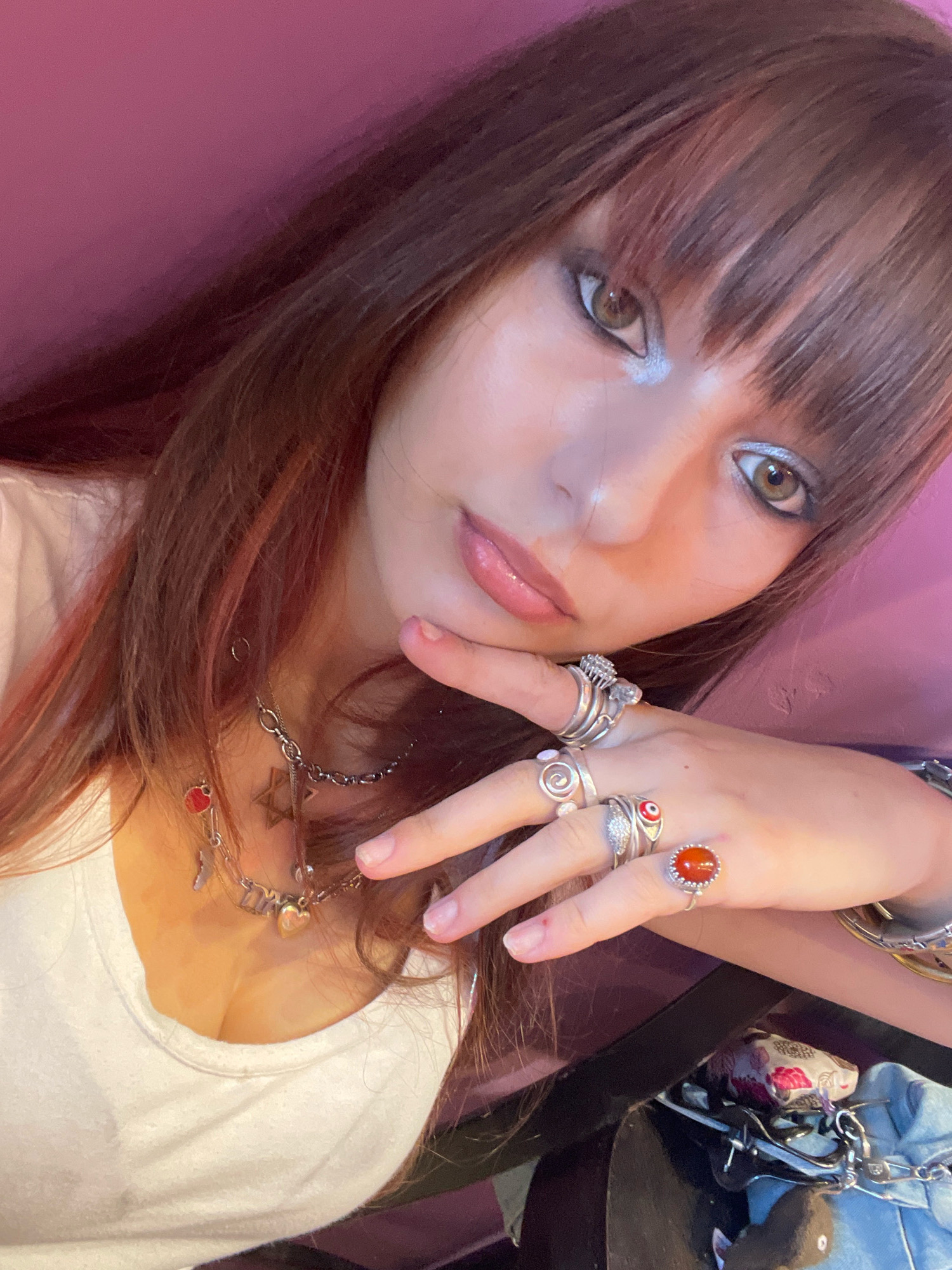 The image shows a close-up, overhead shot of a young woman with long, reddish-brown hair and bangs, wearing a white t-shirt. She is adorned with multiple rings on her fingers, including several silver rings and one with an orange stone, as well as a silver chain necklace with various charms. Her makeup includes silver eyeshadow and eyeliner. She is resting her chin on her hand, with her fingers spread. The background is a solid, deep pink color. The scene is an indoor environment and there is no indication of the city of Raanana, Israel, or any specific urban location.