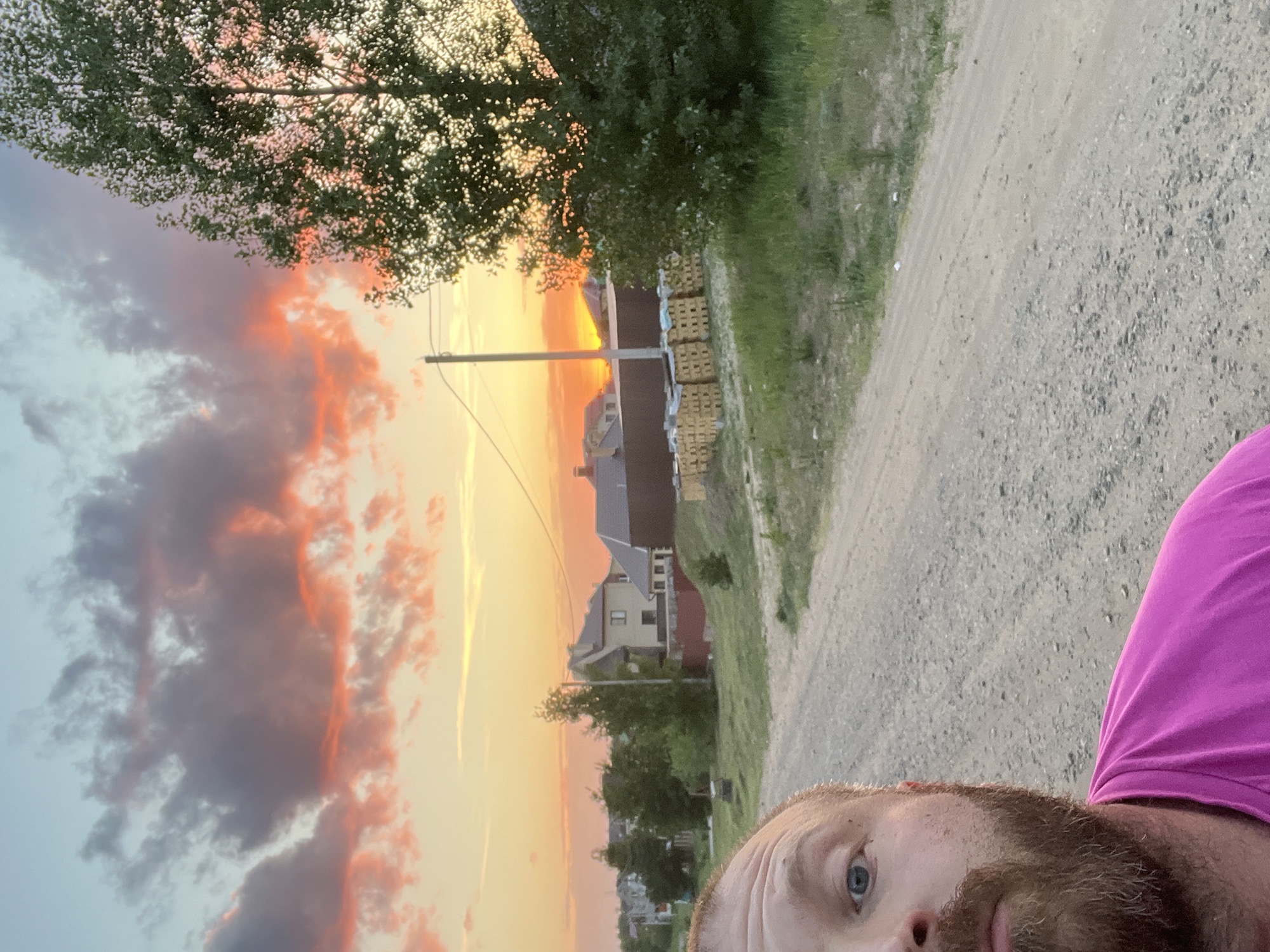 A selfie of a person with a reddish beard, fair skin, and blue eyes is taken in front of a vibrant sunset. The person is wearing a bright pink shirt. The setting appears to be on a gravel road or path. In the background, there are houses, trees, a utility pole, and what appear to be stacks of lumber or materials. The time of day is dusk or dawn, given the warm color of the sky. The approximate location is Noviy, Russia.