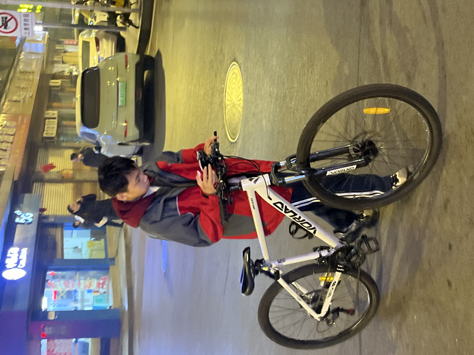 The scene depicts an urban street at night in Jiangjin, China. A male individual, appearing to be in his twenties, stands on a paved surface next to a white VORLAS VR200 mountain bicycle. He is dressed in a red and gray hooded jacket, blue track pants with white stripes, and sneakers. He is actively engaged with a white smartphone, held in both hands, his gaze directed downwards.
The bicycle features a white frame with "VORLAS" branding, black handlebars, saddle, and tires, front suspension forks, disc brakes on both wheels, and a yellow reflector attached to the front wheel spokes. A bottle cage is affixed to the frame's downtube.
In the background to the left, a brightly illuminated storefront displays the Chinese characters for "China Mobile" (中国移动) and "5G". Several indistinct individuals are visible inside or near the store, one appearing to use a phone. To the right, a light green electric vehicle with a distinctive green Chinese license plate is visible on the street. Further beyond, other vehicles and a "no left turn" traffic sign are partially discernible. The area is illuminated by artificial streetlights, casting a yellowish glow on the pavement, where a circular manhole cover with a decorative pattern is present.