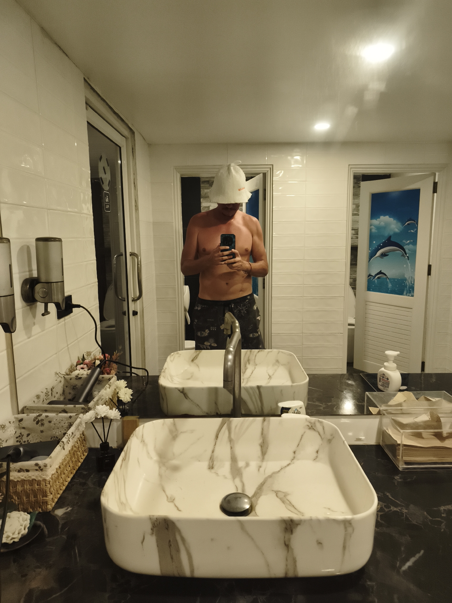 A man is taking a photo of himself in a bathroom. He is wearing a white sauna hat and swimming trunks. The bathroom features marble sinks, white tile walls, and two doors, one with a dolphin image. The location is บ้านบางรักษ์, Thailand.