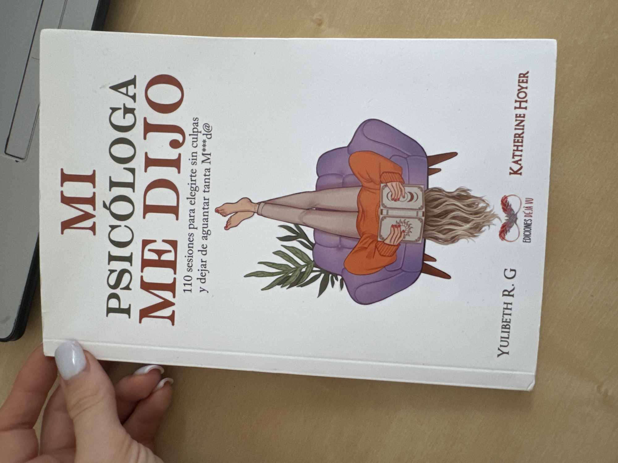 A book cover with the title "MI PSICÓLOGA ME DIJO" is displayed on a wooden surface. The book is white with red and dark gray text. The title is large and centered at the top. Below the title, there is a subtitle that reads "110 sesiones para elegirte sin culpas y dejar de aguantar tanta M***d@". The cover features an illustration of a person lying on a purple armchair with their legs in the air, reading a book. The person is wearing an orange sweater and beige pants. The bottom of the book shows the names "YULIBETH R. G", "EDICIONES DÉJÀ VU", and "KATHERINE HOYER". The image does not contain any information about the city of Murcia, Spain.