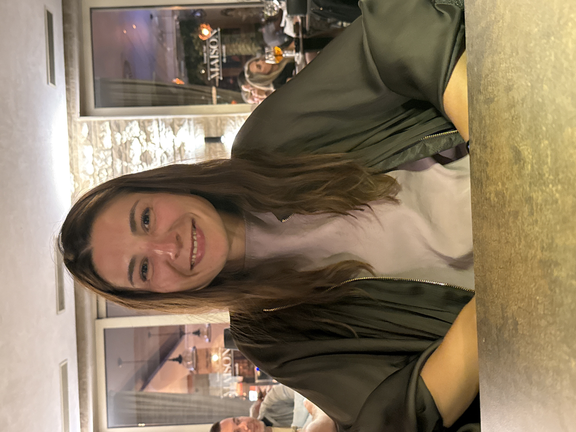 A medium shot depicts a woman seated at a table, smiling at the camera. She has brown hair and is wearing a light purple shirt and a dark green jacket. In the background, a window reflects the exterior of a building with the word "MAISON" visible. Other individuals are visible in the background, both in the reflection and directly behind the woman. The table has a marbled surface. Based on the reflections in the window and the general ambiance, the location appears to be a restaurant or similar establishment. The city of Đenovići, Montenegro is not evident from the visual elements alone, the image is primarily an interior shot.