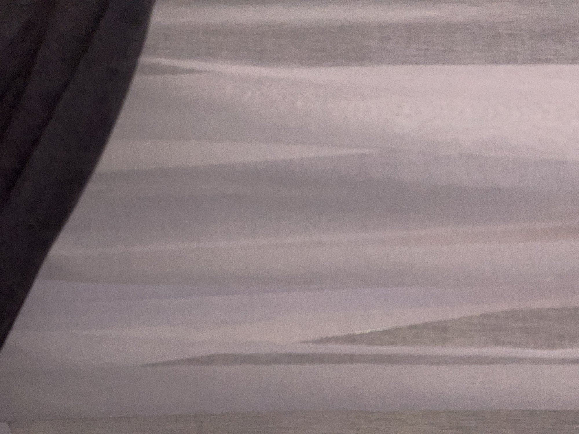 The image shows a close-up of draped fabric, likely curtains or a soft furnishing. The fabric is a light, translucent material, possibly sheer or semi-sheer, in shades of white and light pink, with vertical folds and pleats that create subtle shadows and highlights. A darker, opaque material, possibly black or dark gray, is visible along the top edge of the frame, casting a shadow onto the lighter fabric below. The texture of the fabric appears soft and slightly textured, with some visible grain or speckling, possibly due to the lighting or the material itself. There are no people, animals, or specific objects clearly identifiable beyond the fabric. The background is indistinct, suggesting a shallow depth of field or the nature of the material obscuring whatever is behind it. The scene is static and appears to be indoors. The provided location information "Doma 1096 Km, Russia" is not visually derivable from the image itself.
