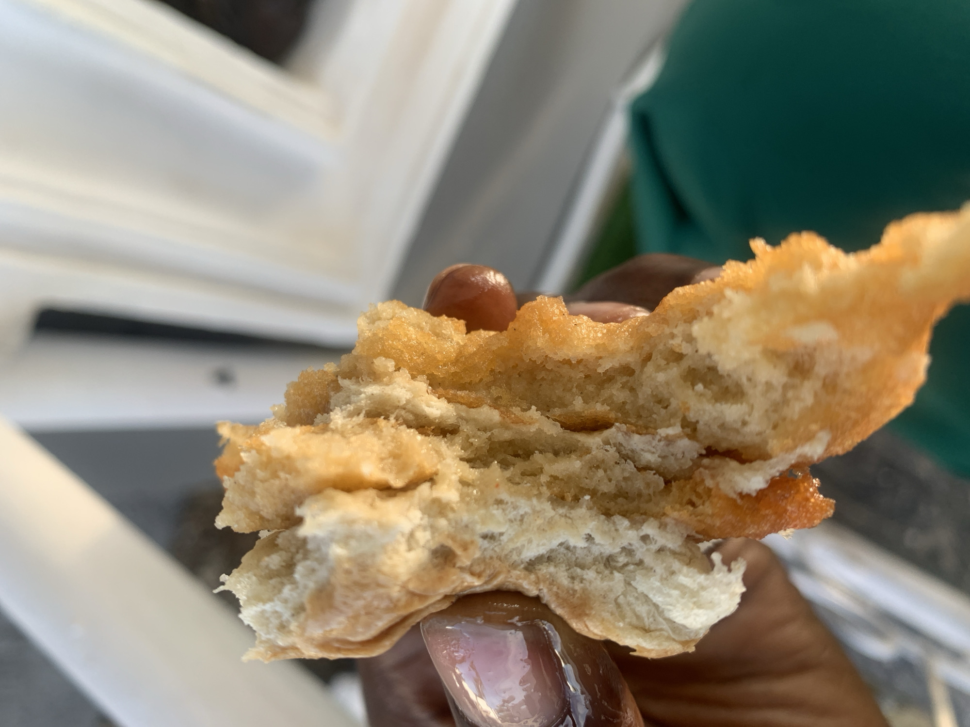 A close-up shot shows a person's hand holding a piece of fried dough, possibly puff puff, with a visible orange filling, being broken in half. The background is blurred, but suggests an outdoor setting with white vertical structures and a hint of green. The lighting indicates it is daytime. The location is Gura Top, Nigeria.