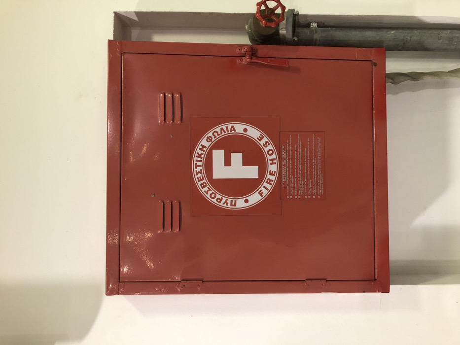 The image displays a red, rectangular metal fire hose cabinet mounted on a white wall in what appears to be an indoor setting in Moschato, Greece. The cabinet is positioned centrally in the frame, with white wall space visible to its left and below. Above the cabinet, a gray metal pipe with a red valve is visible, suggesting a water supply line for the fire hose.
The cabinet door features a prominent white circular logo in the center. The logo contains a large, stylized white letter "F" in its middle. Around the "F," written in white text within the circle, are the Greek words "ΠΥΡΟΣΒΕΣΤΙΚΗ ΦΩΛΙΑ" (Pyrosvestiki Folia), which translates to "Fire Hose Cabinet" or "Firefighting Nest," and the English words "FIRE HOSE."
To the right of the circular logo, a smaller rectangular label with white text on a darker background (likely red) is affixed. This label appears to contain instructions or regulations, predominantly in Greek, with several bullet points.
The cabinet door has three vertical indented lines (vents or decorative elements) on both its upper left and lower left sections. The top of the cabinet door is secured by two visible hinges, and a red metal latch handle is positioned near the top center, allowing the door to be opened. The metal surface of the cabinet shows some minor wear and dust. Shadows are cast by the cabinet onto the white wall, indicating an overhead light source.
