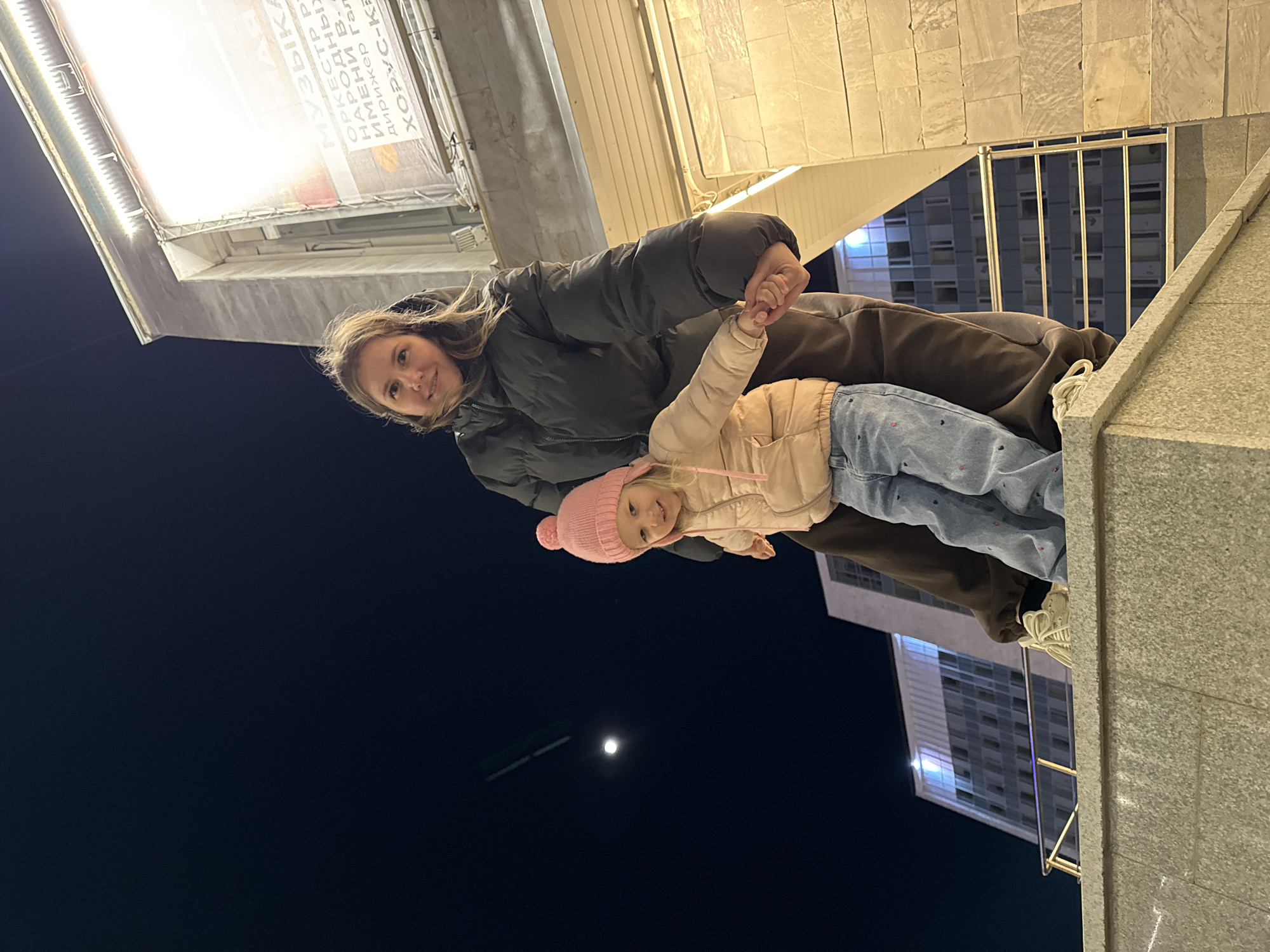 A mother and her daughter are standing on a concrete ledge at night. The mother is wearing a dark puffer jacket and brown pants. She is holding her daughter's hand. The daughter is wearing a pink hat and jacket, and blue jeans. They are standing in front of a building with a sign in Cyrillic. In the dark sky, a bright moon is visible. The location is Perm, Russia.