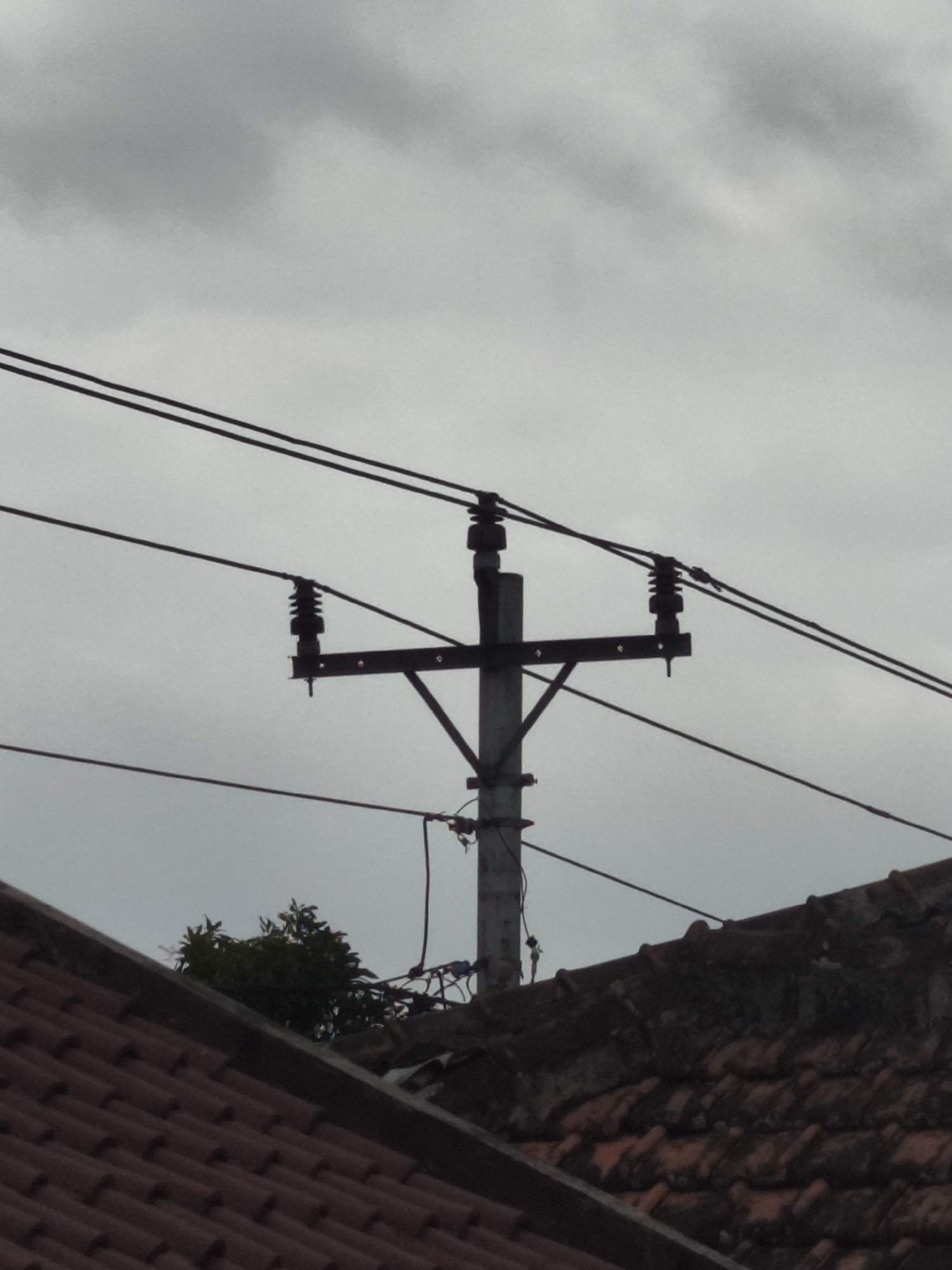 The image depicts a utility pole with electrical wires against a cloudy sky, situated in the city of Yogyakarta, Indonesia. The pole is made of concrete and has crossbars supporting the overhead lines, which are insulators. In the foreground, a portion of a tiled roof is visible, suggesting it is part of a residential or commercial building. The scene is set outdoors, under overcast conditions, with the cloudy sky dominating the upper portion of the frame.