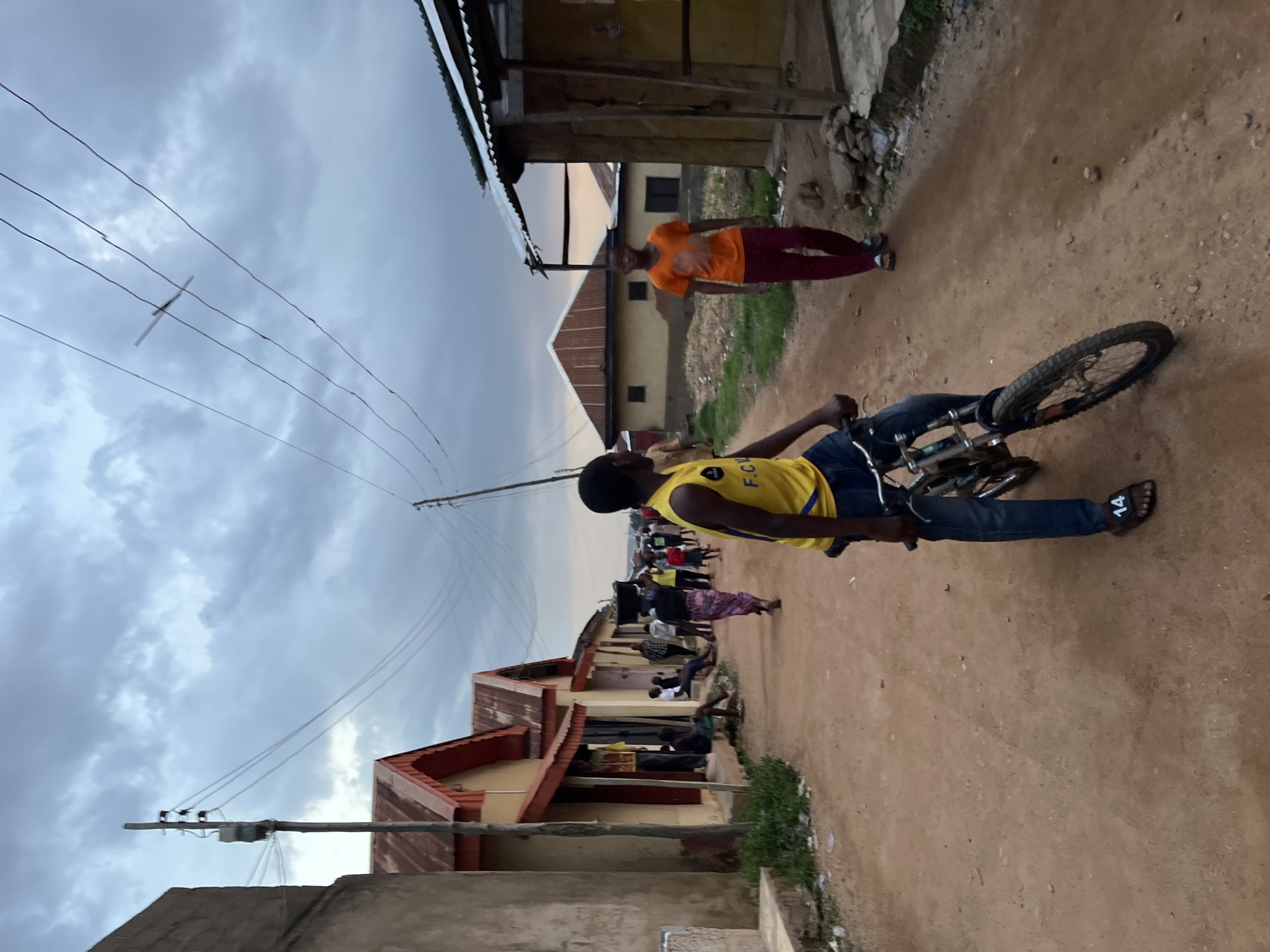 A group of people walk down a dirt road in the city of Jos, Nigeria. In the foreground, a boy in a yellow shirt and blue jeans is on a bicycle. Behind him, a person in an orange shirt and red pants walks down the road. Further in the background, several people are walking, and some are sitting outside buildings. The buildings are simple structures with corrugated metal roofs. The sky is overcast, suggesting it is late in the day or there is cloud cover. Utility poles and wires are visible, indicating the presence of electricity infrastructure. The overall scene depicts a typical street in a residential area.