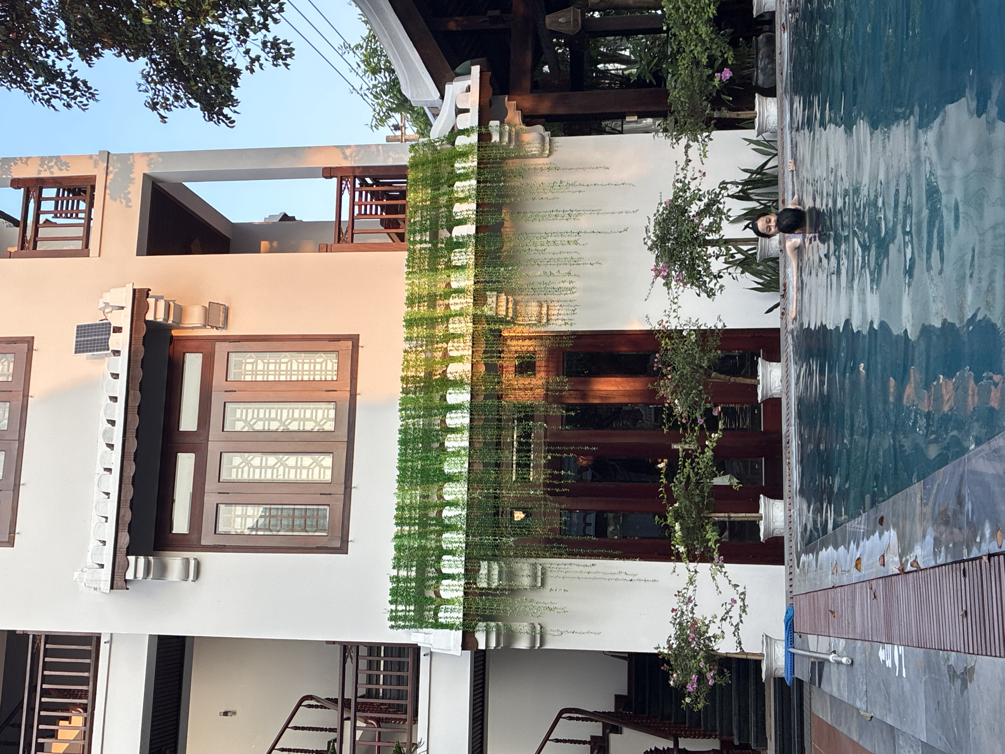 The image shows a two-story building with white walls and dark wood trim next to a swimming pool. The building has several windows with intricate designs. Hanging plants adorn the building's facade. A person is in the pool. The location is in Hội An Ward, Vietnam.